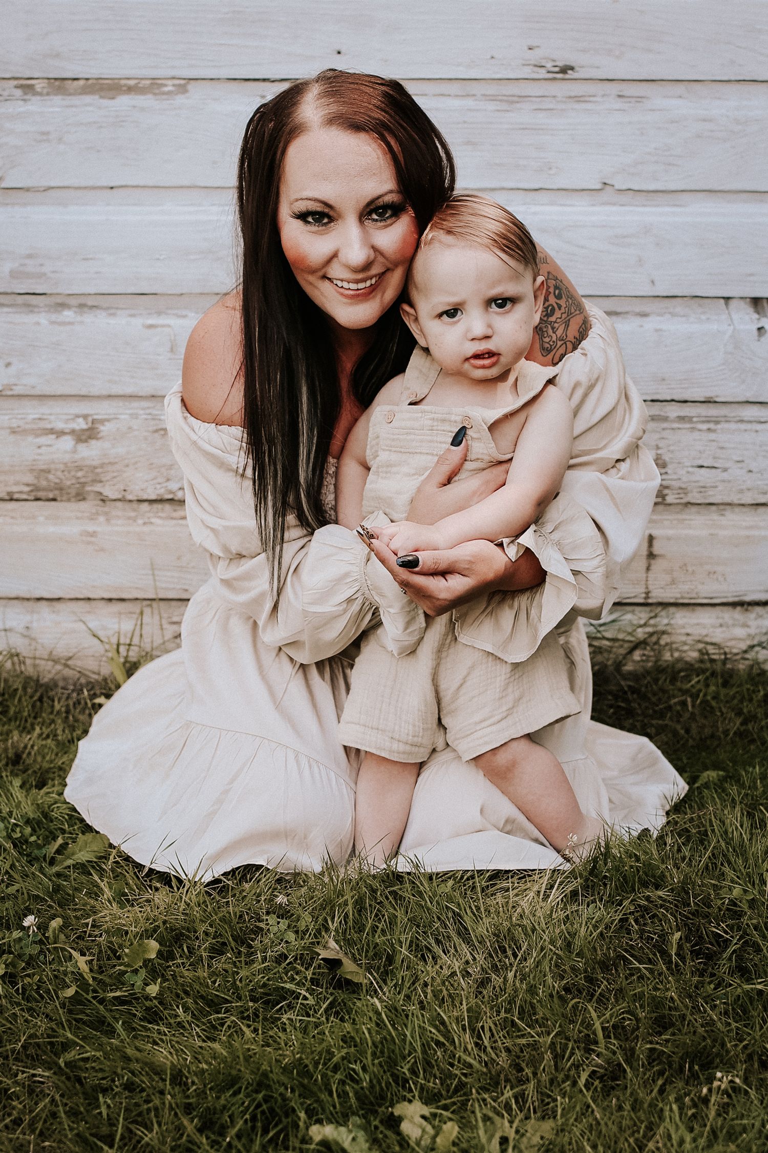 A parent and child wearing matching cream-colored dresses sit together on grass against a rustic wooden wall.