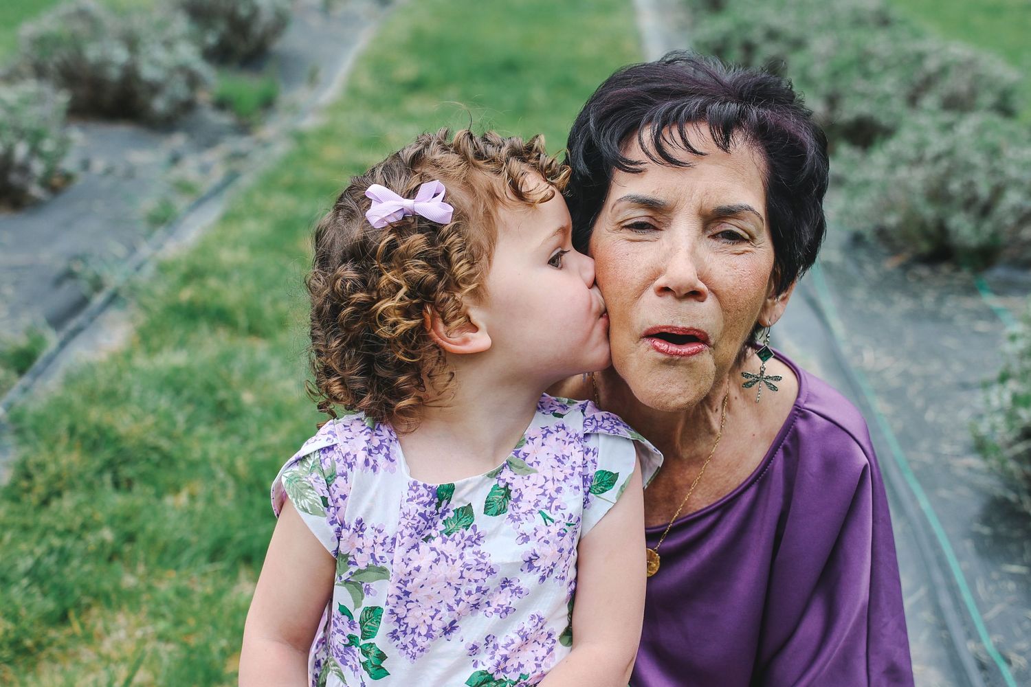 A young child with curly hair wearing a floral dress gives a sweet kiss on the cheek outdoors in a garden setting.