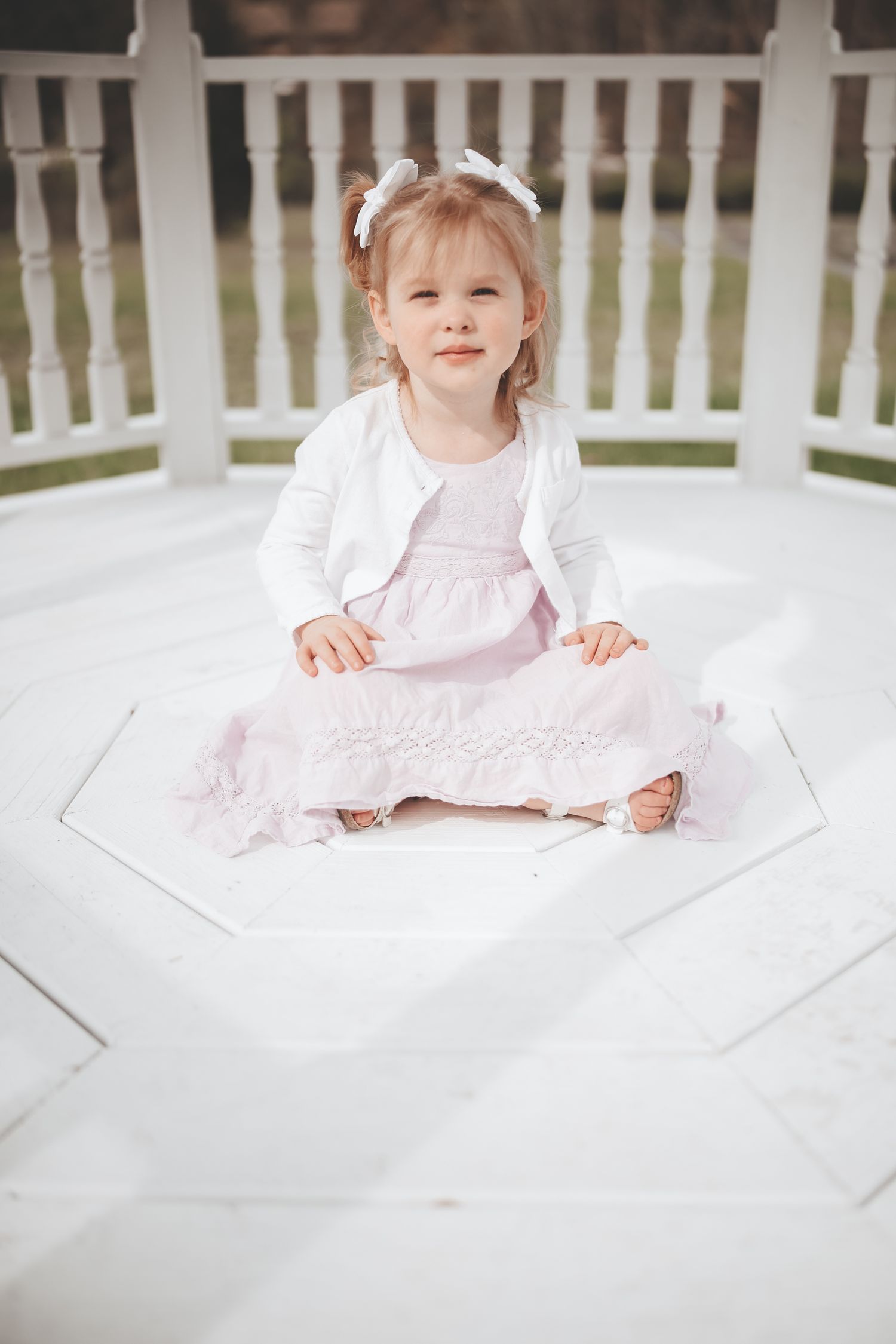 A small figure in a white dress sits on a white porch with a picket fence in the background.