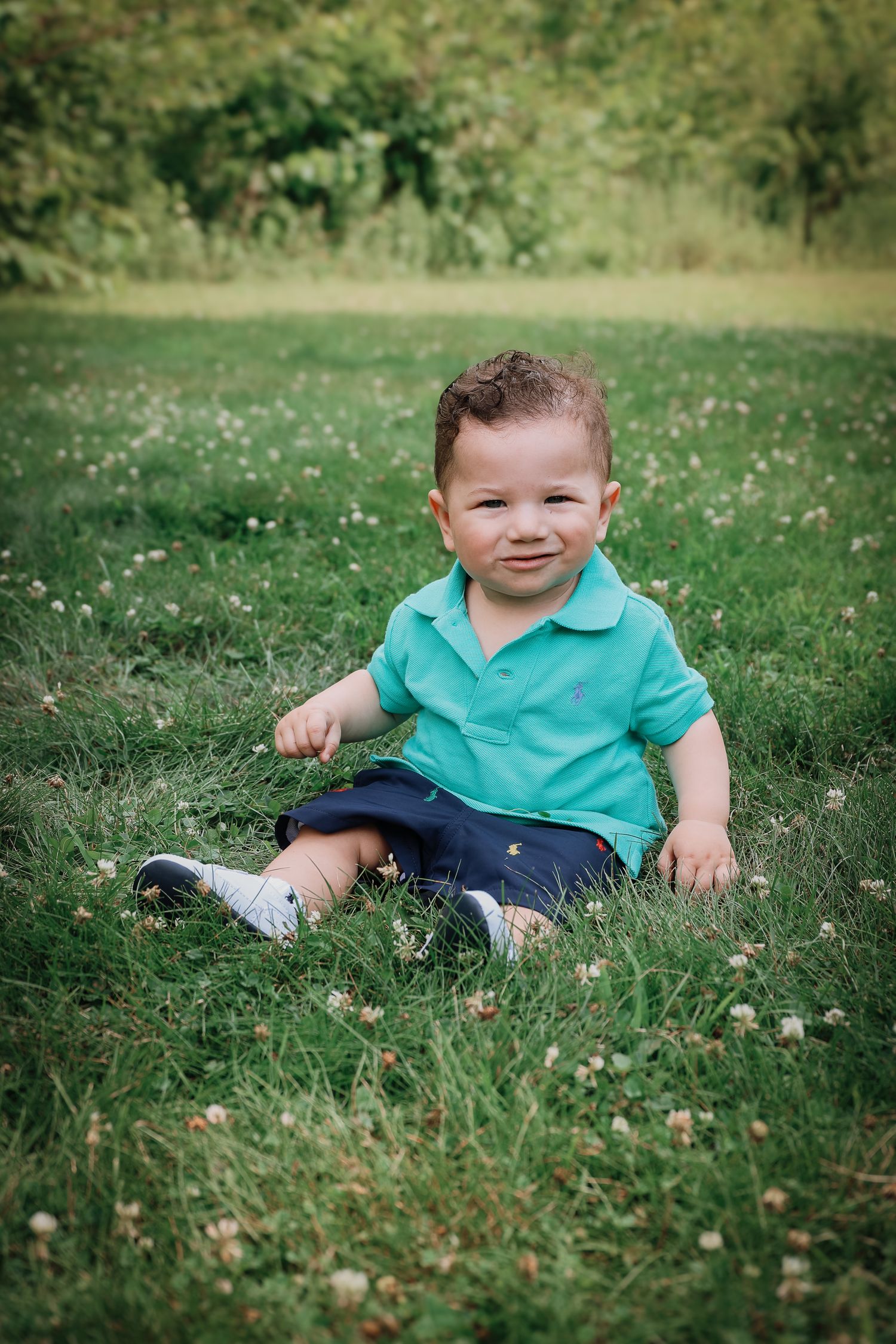 A small child in a turquoise polo shirt and navy shorts sits in a grassy field with white wildflowers.