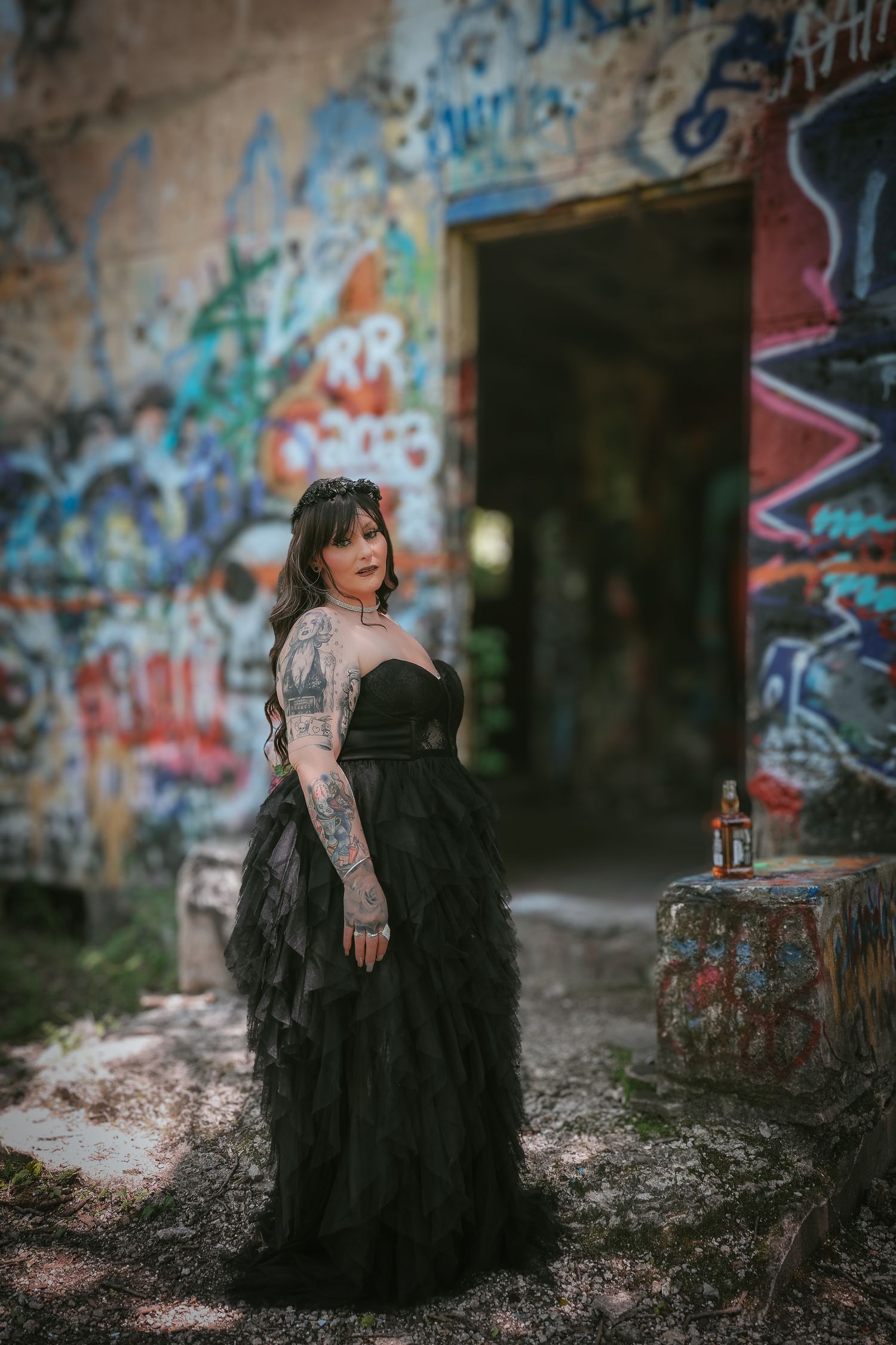A figure in a flowing black dress poses dramatically against a graffiti-covered wall in an urban setting.