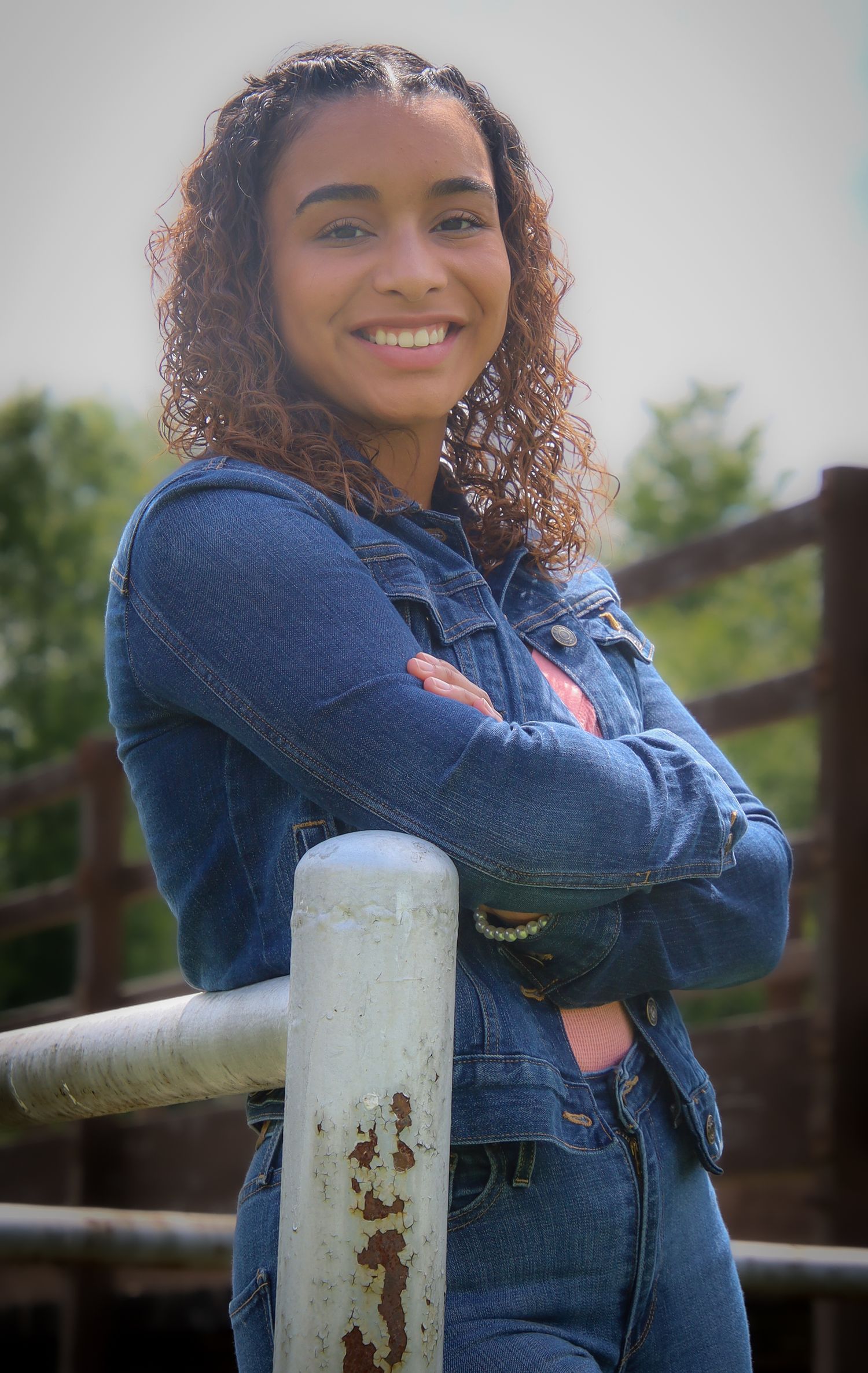 A person in a denim jacket leans against a rustic white railing while posing outdoors with natural lighting.