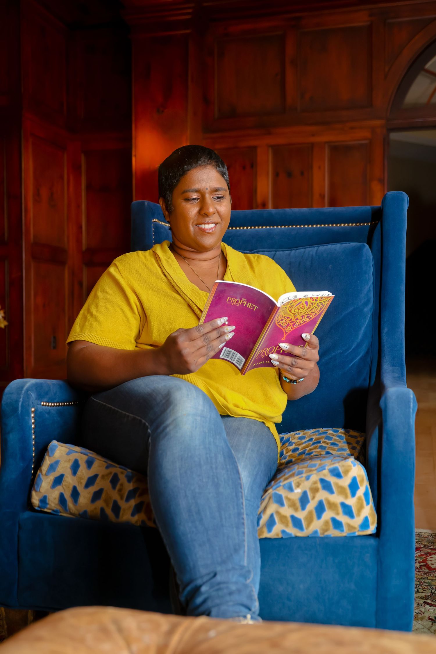 Someone in a yellow shirt and jeans sits comfortably on a blue chair while reading a small pink book.