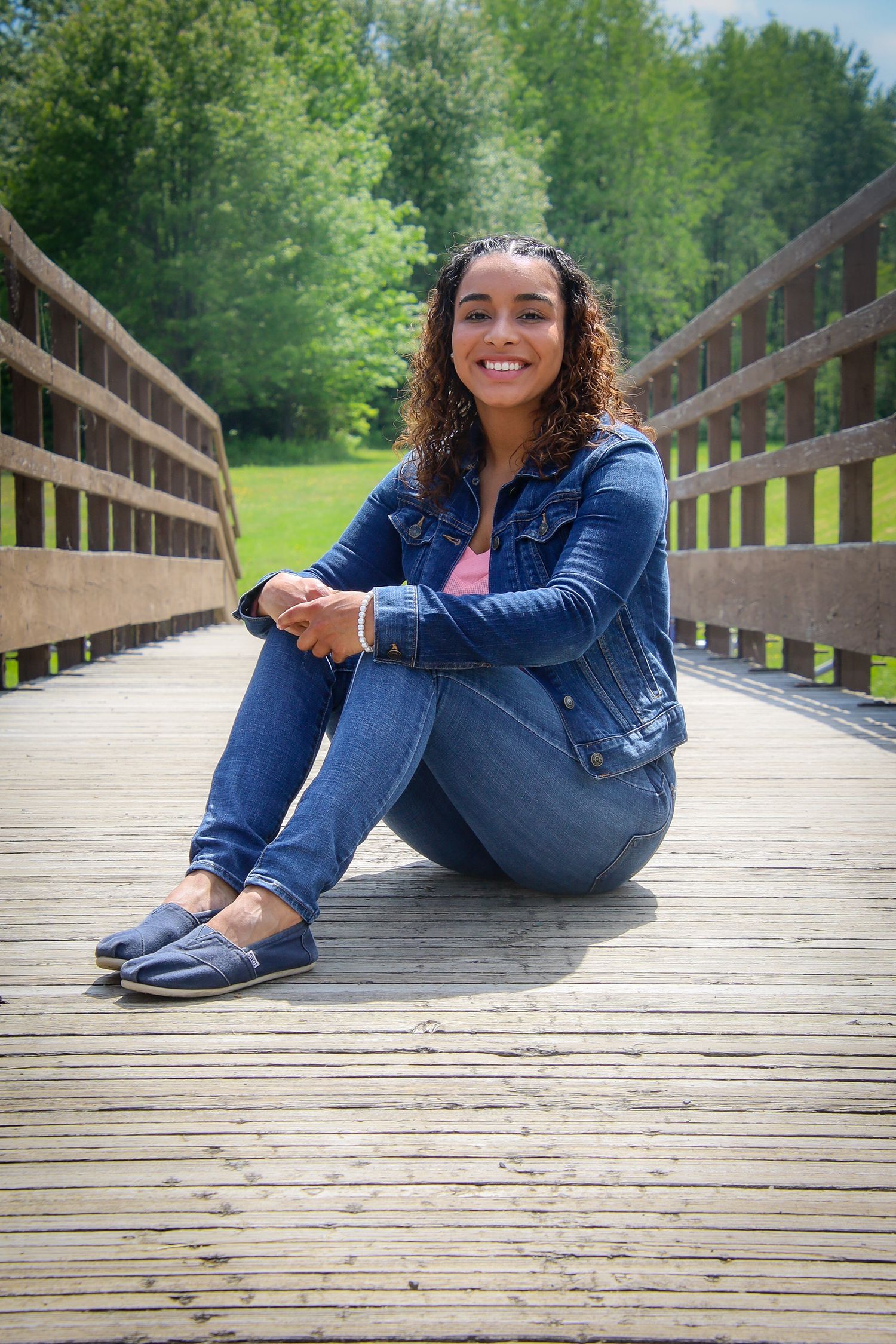 Person in denim sitting casually on wooden boardwalk surrounded by lush green trees on sunny day.