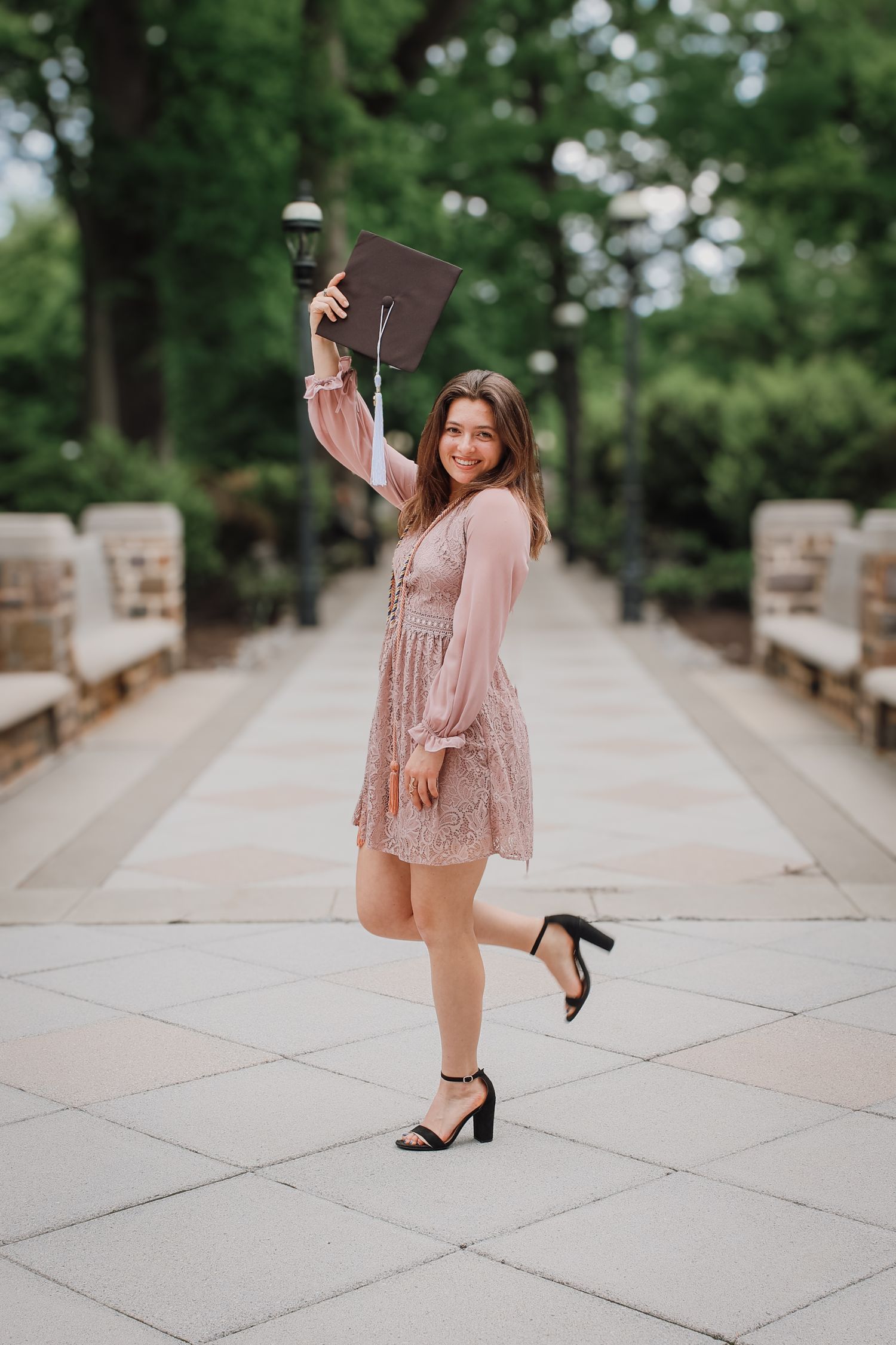 A graduate in a sparkly pink dress and cap poses playfully on a concrete walkway during a graduation photoshoot.