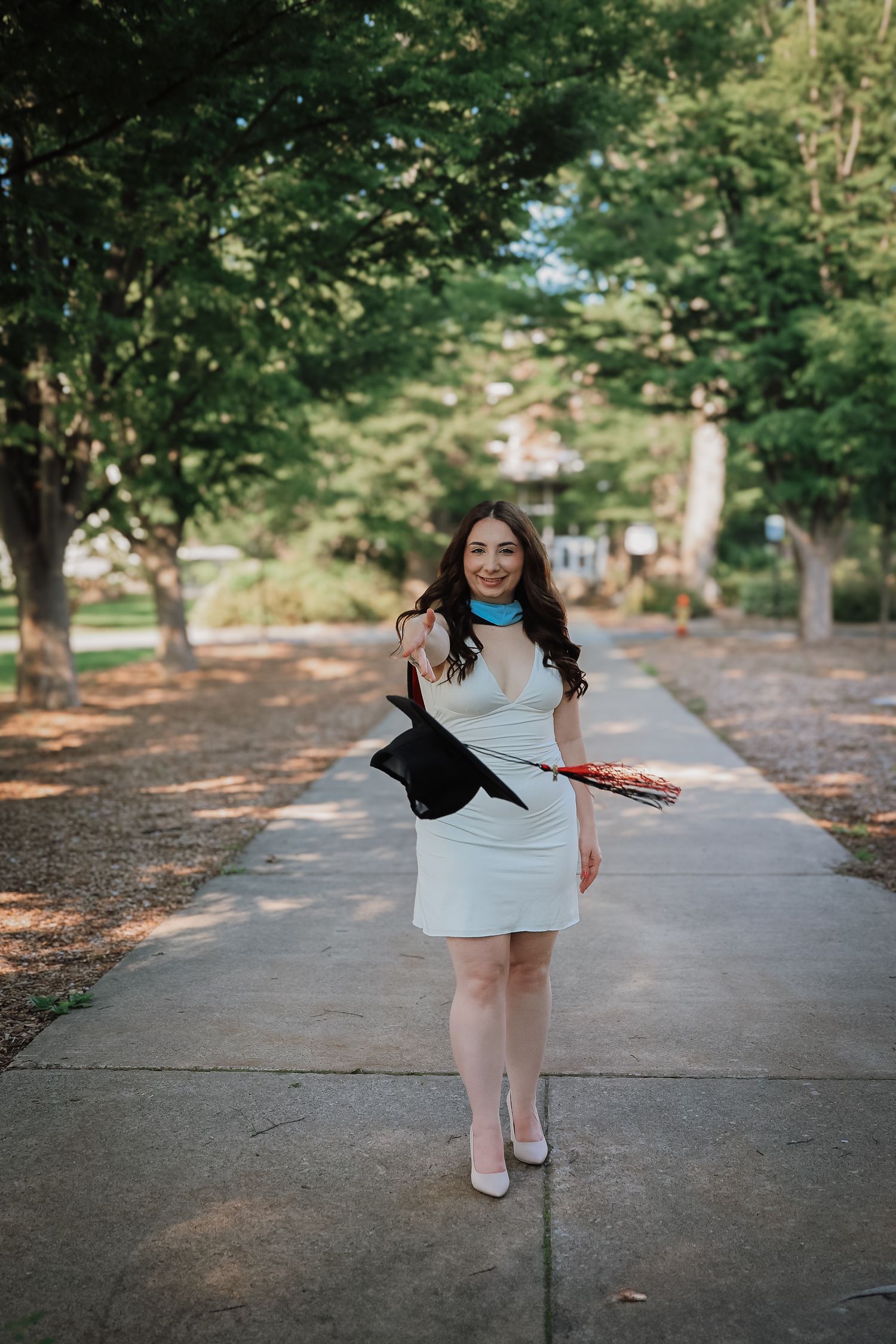 A person in a white dress twirls and dances playfully on a tree-lined path during a sunny afternoon.