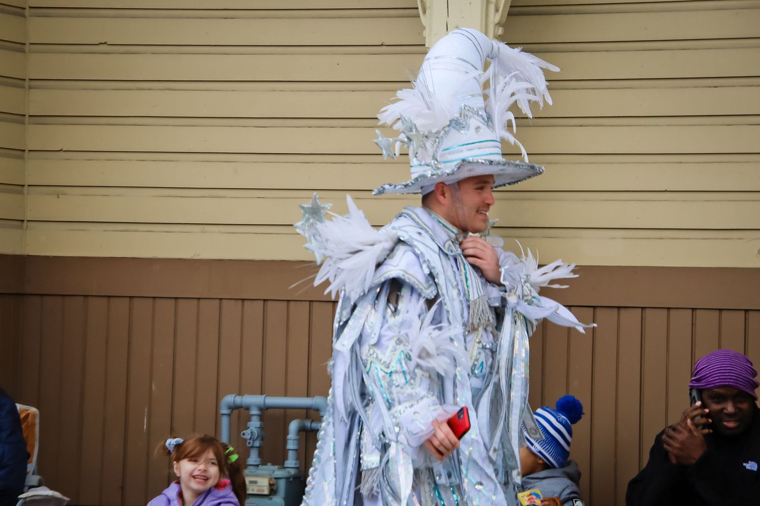 A street performer dressed in an elaborate white and silver costume with feathers stands against a beige building exterior.