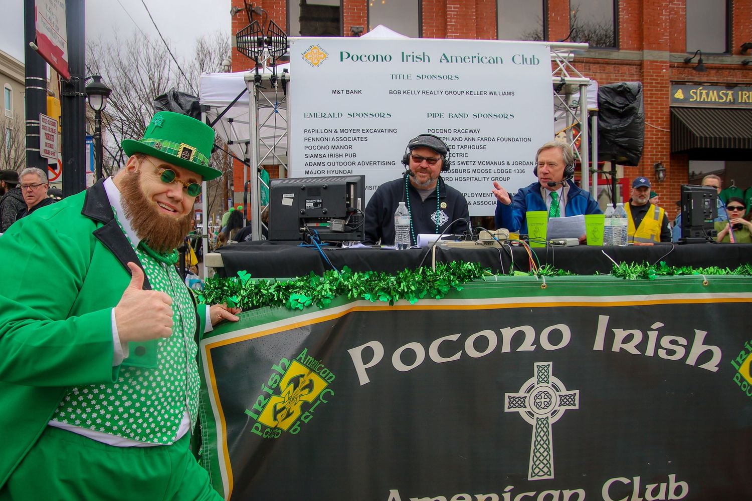 A person in bright green St. Patrick's Day attire stands by the Pocono Irish American Club banner at a festive event.