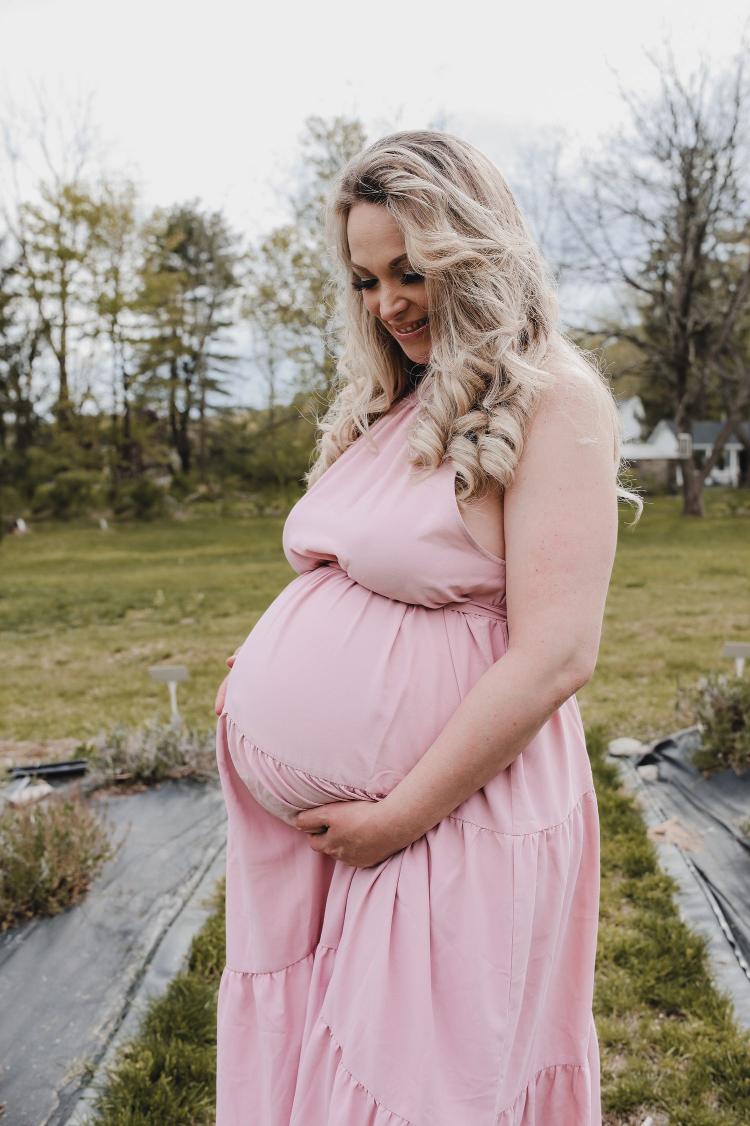 A pregnant person in a flowing pink dress stands outdoors in a grassy field on a sunny day.
