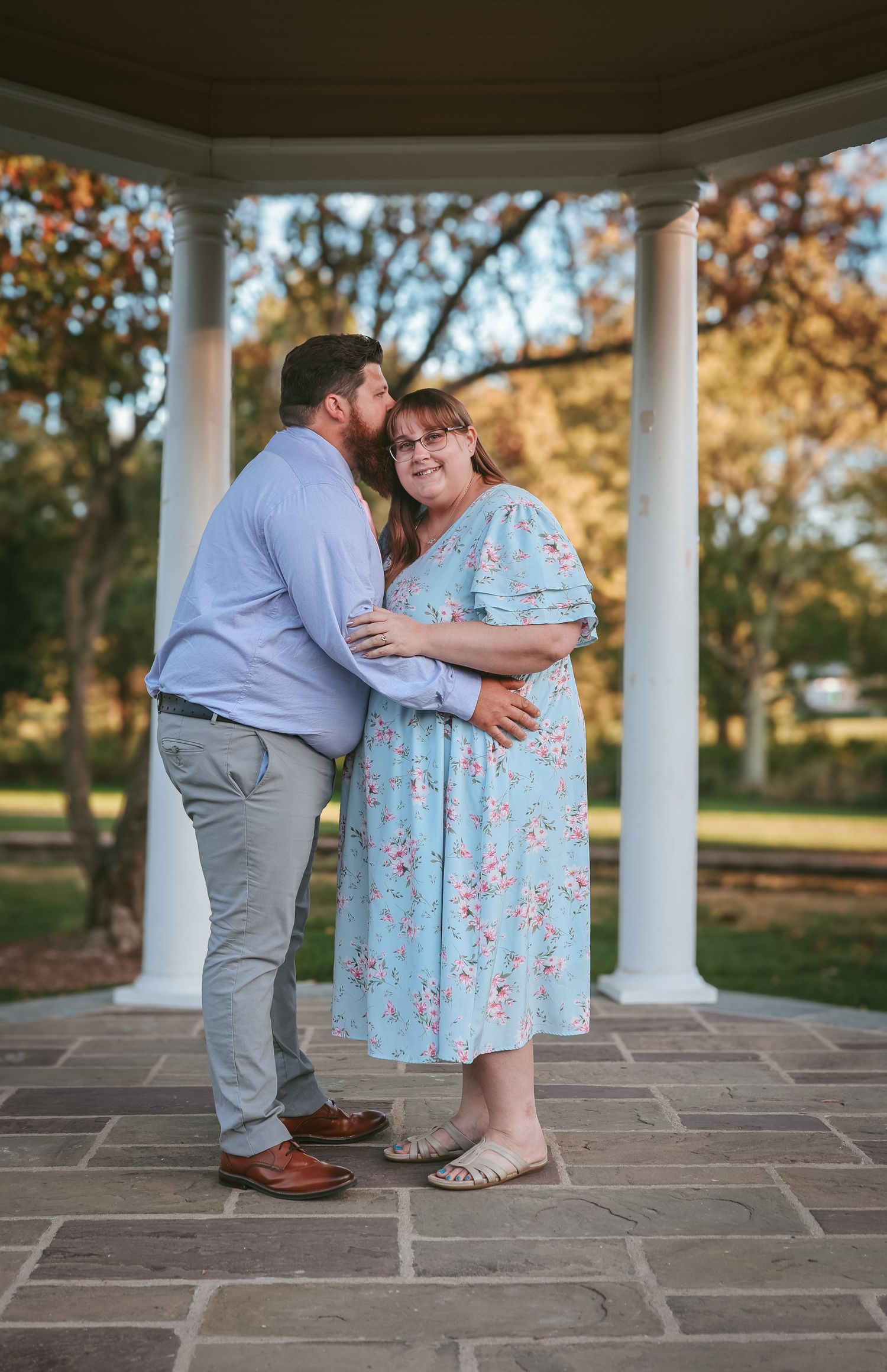A couple shares a tender moment in a white columned gazebo during golden hour on stone pavers.
