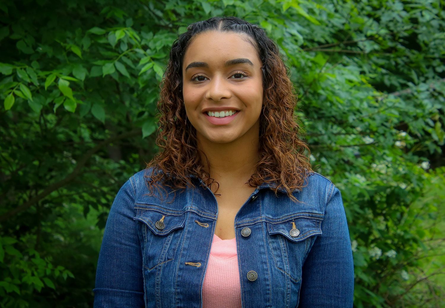 A person with curly brown hair wearing a denim jacket and pink top smiles while standing in front of green foliage.