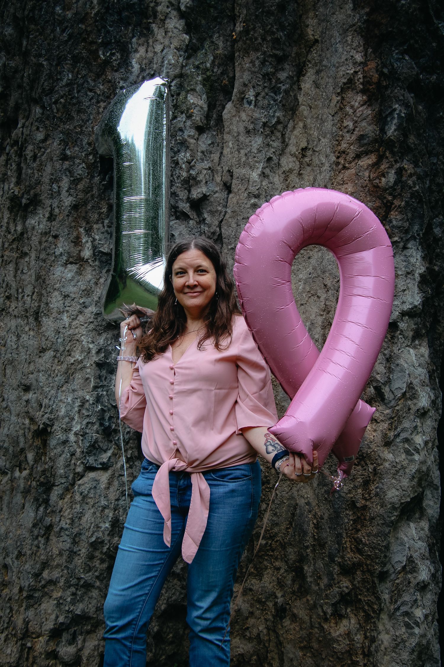 Person in pink button-up shirt and jeans holds pink inflatable number while standing in front of waterfall.