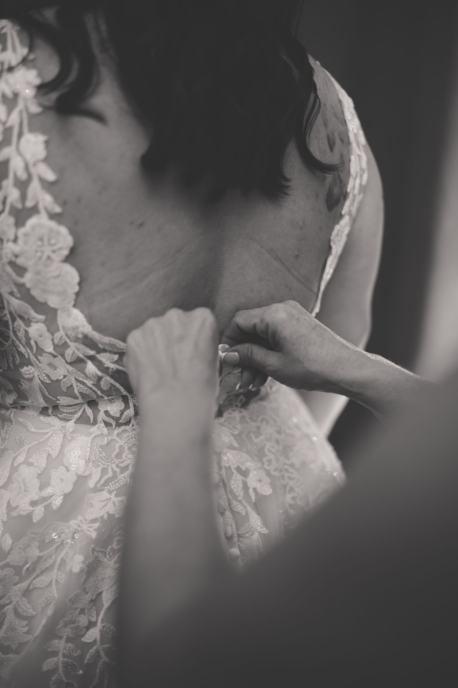 Close-up black and white photo showing hands fastening delicate lace details on a wedding dress.