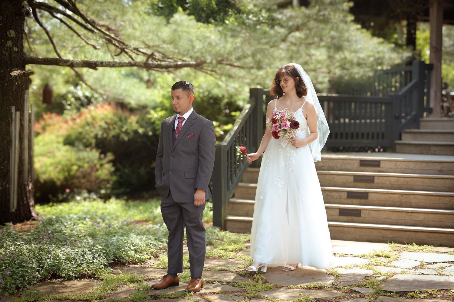 A wedding couple and their wedding party pose outdoors in a garden setting on a sunny day.