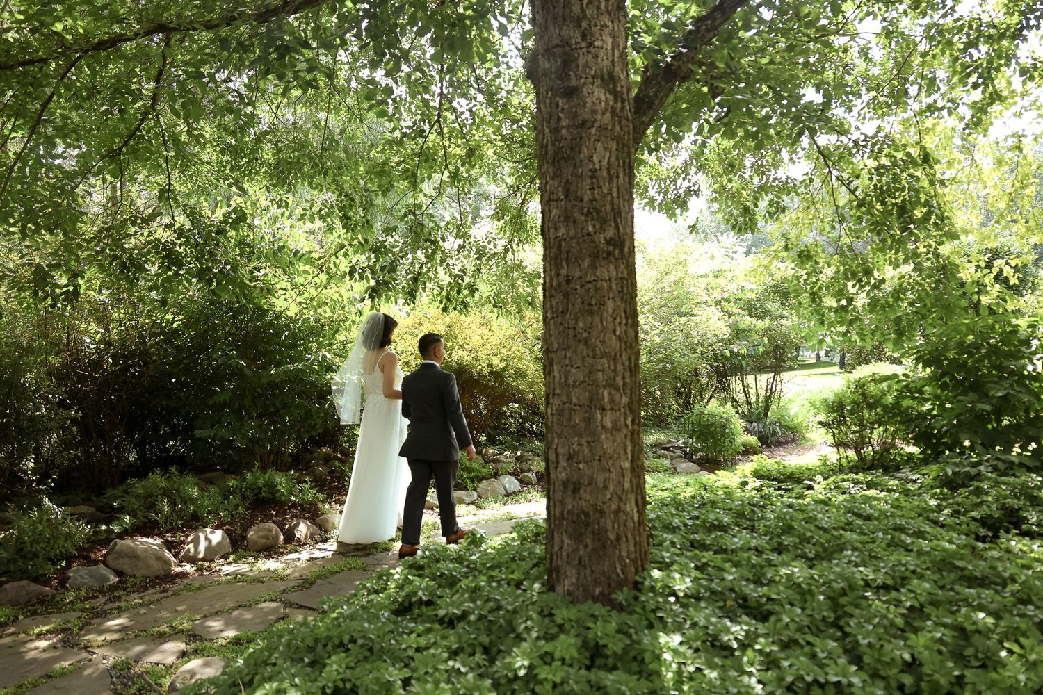 Two figures in wedding attire stand together beneath a large tree in a lush garden setting.