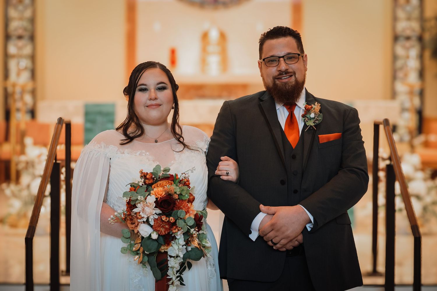 A wedding party walks down a church aisle with orange and peach floral bouquets and decorative accents.