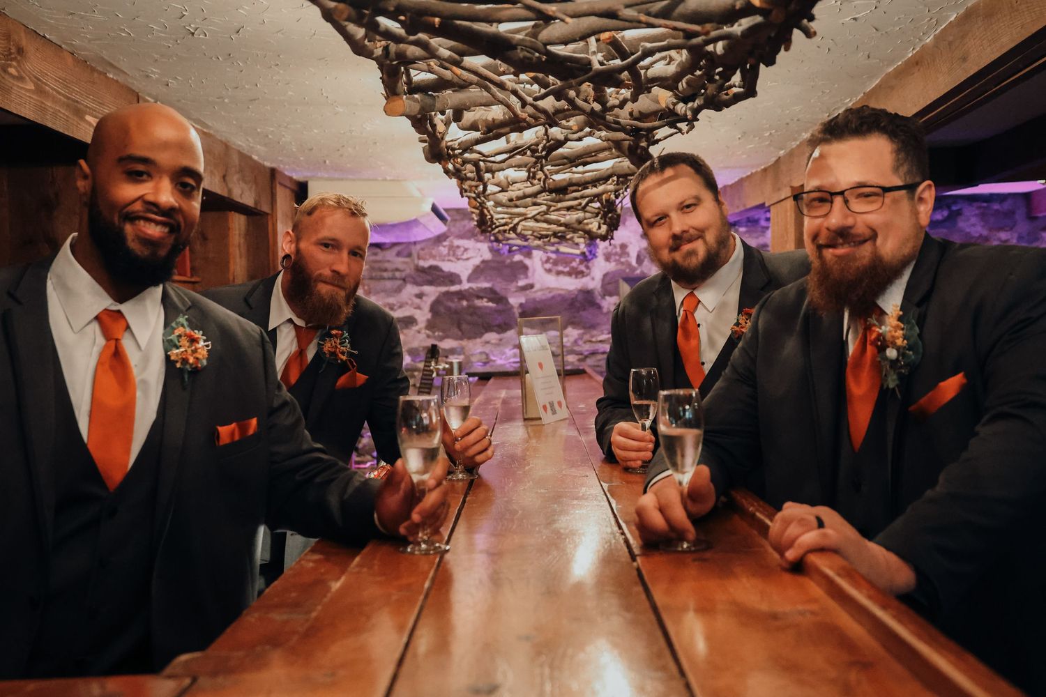 A group of groomsmen in dark suits and orange ties pose together at a wedding celebration.