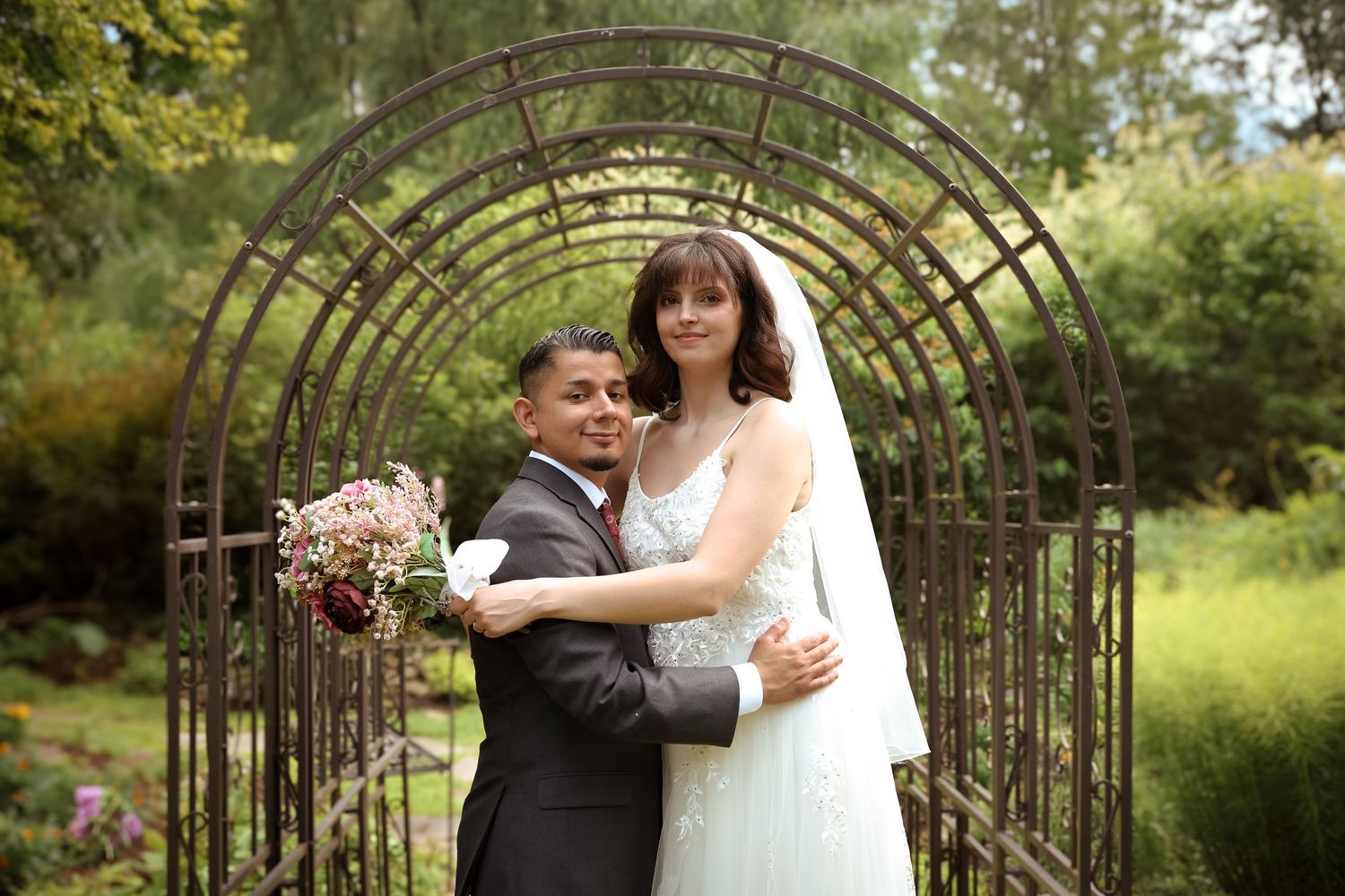 A couple poses under a rustic metal garden arch during their wedding ceremony in a lush outdoor setting.