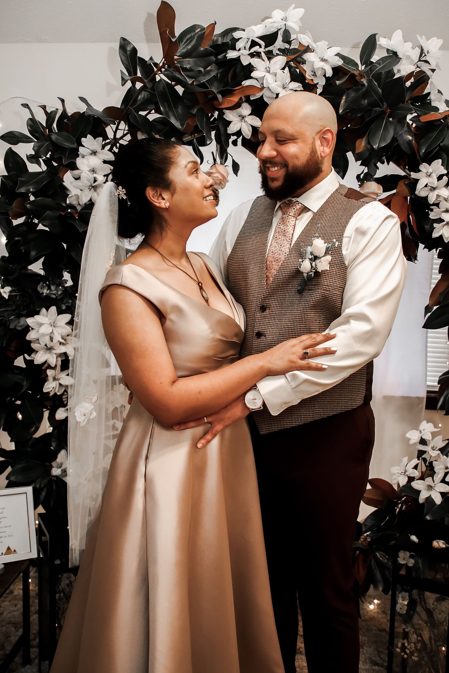 A couple shares a joyful moment under a floral arch with white flowers and dark foliage at their wedding ceremony.