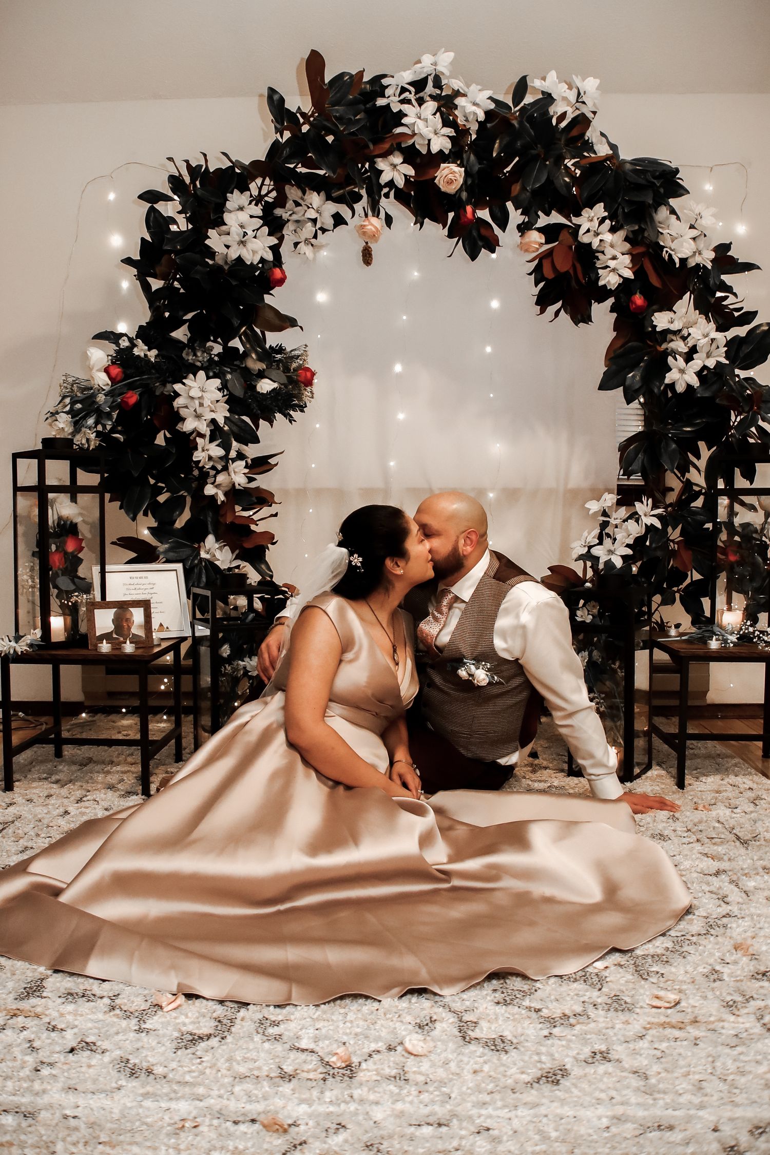 A couple in formal attire lies on a white carpet under floral decorations during an intimate photo session.