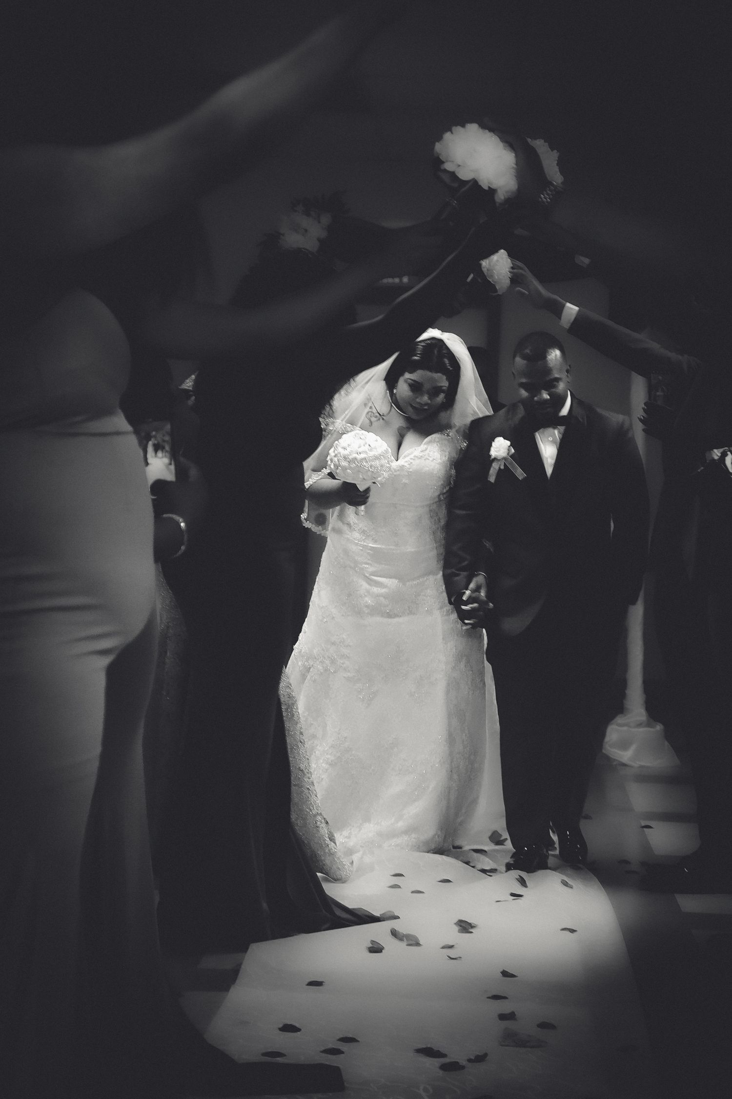 A couple in formal wedding attire dances together in a dimly lit ballroom with scattered petals on the floor.