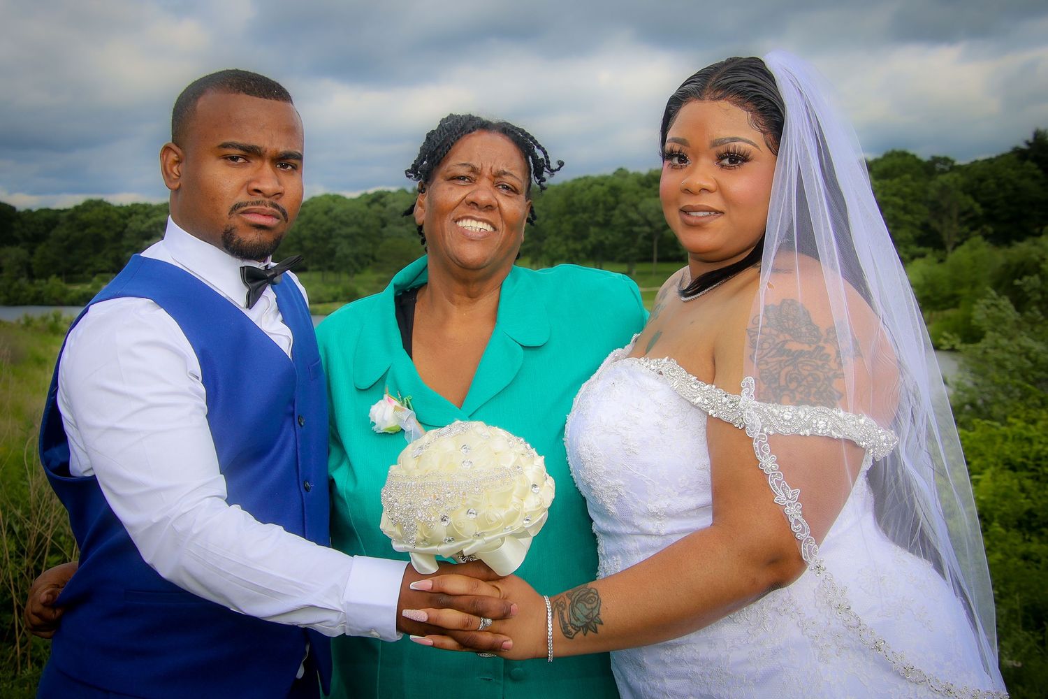 Three people pose together outdoors at a wedding celebration with a white bouquet against a green landscape background.