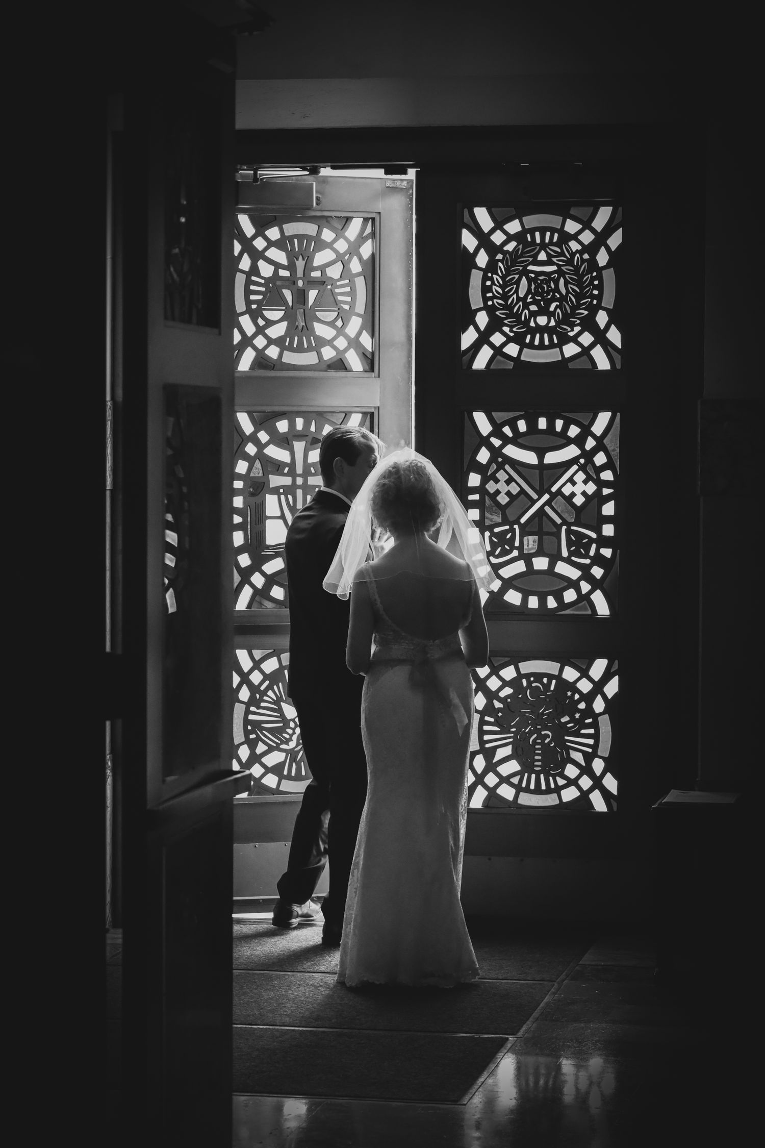 A black and white silhouette of a bride in a wedding dress standing before ornate stained glass windows.
