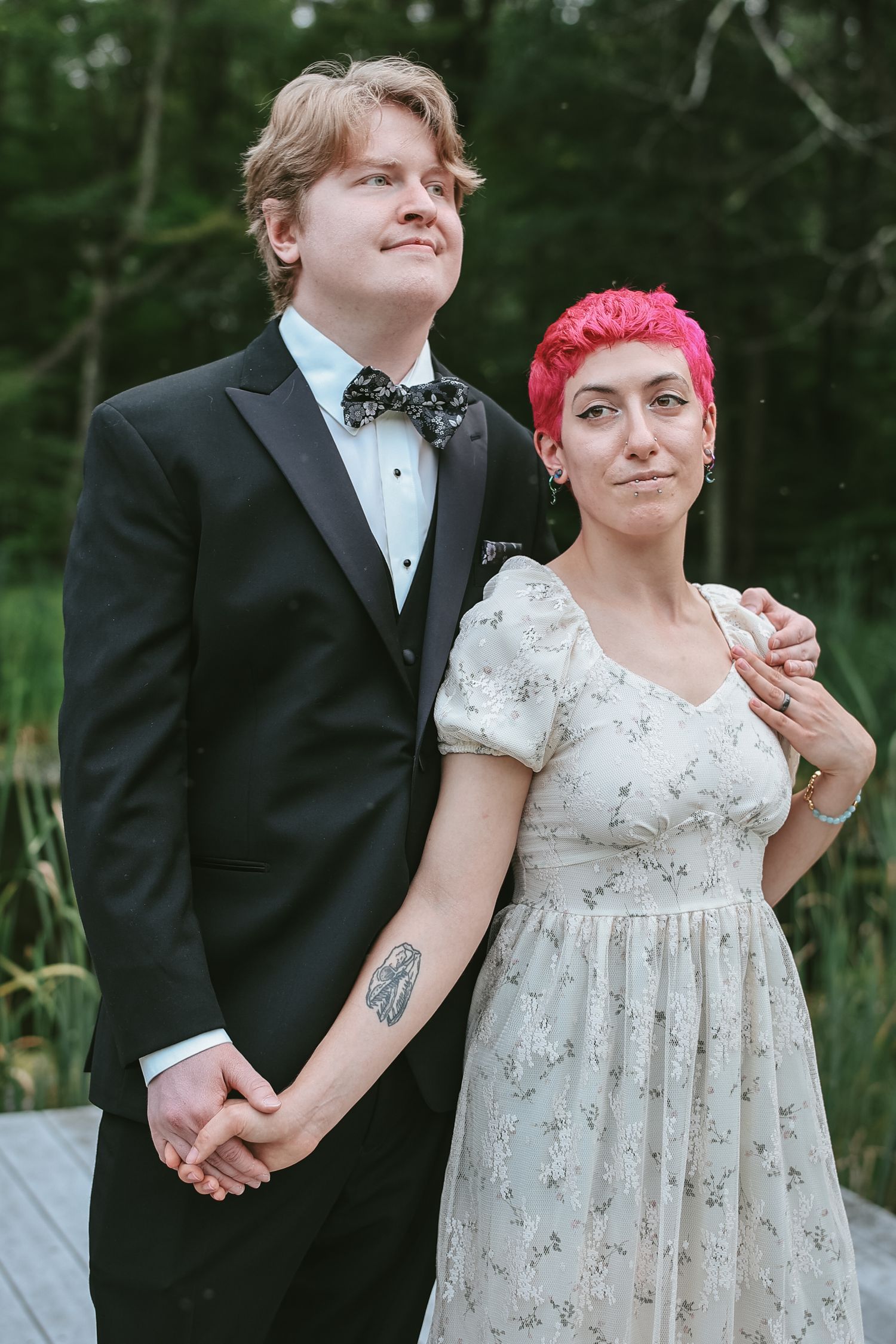 A couple in formal wedding attire poses outdoors in a garden setting with trees in the background.