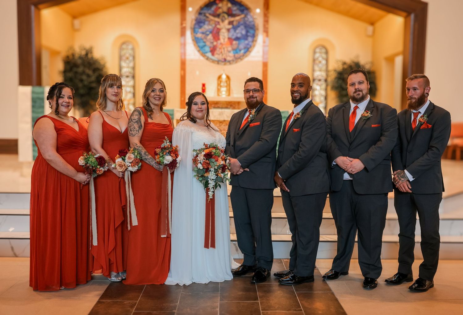 Wedding party members stand in a row wearing red dresses and dark suits inside an elegant church venue.