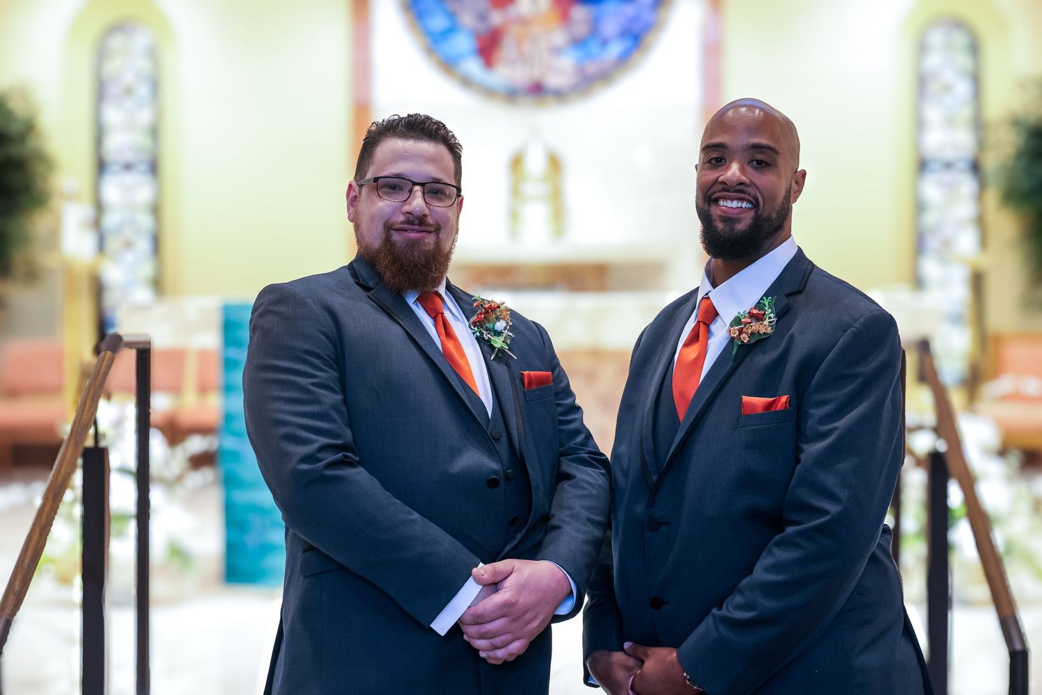 Two groomsmen in black suits with red accents stand together at the altar of a church during a wedding ceremony.