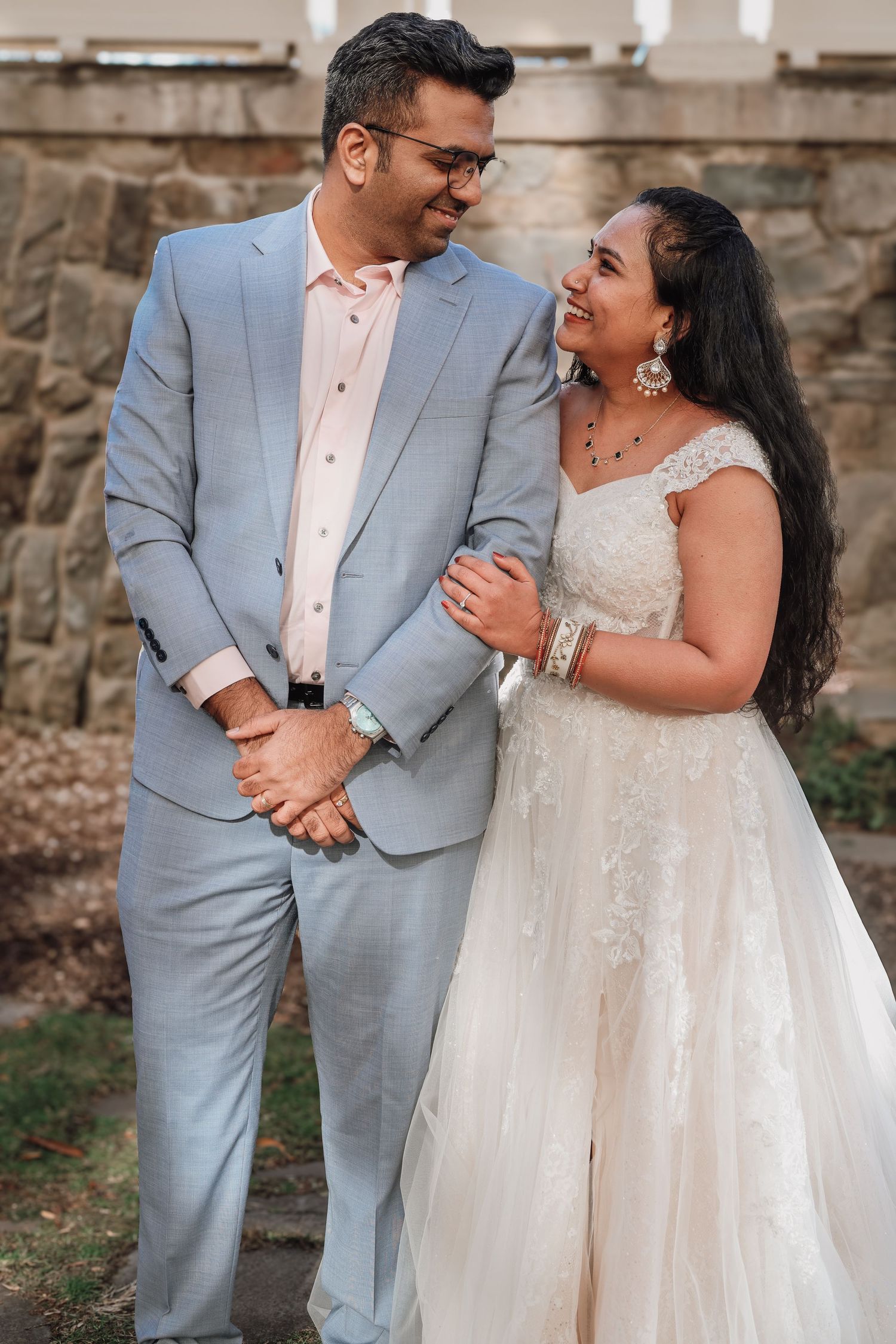 A romantic wedding couple shares intimate moments in front of a stone wall wearing a light gray suit and white lace gown.