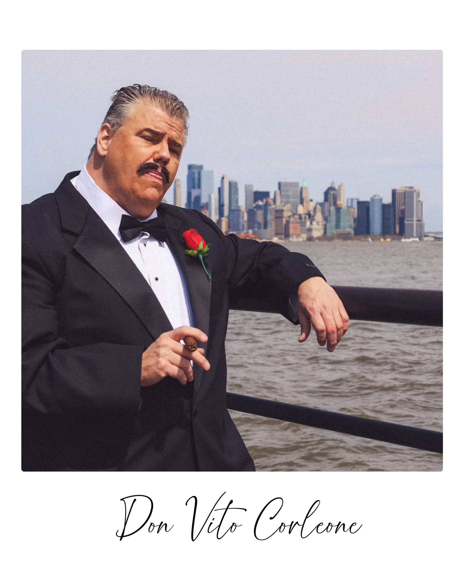 Person in formal black tuxedo and red boutonniere stands against waterfront cityscape backdrop at dusk.