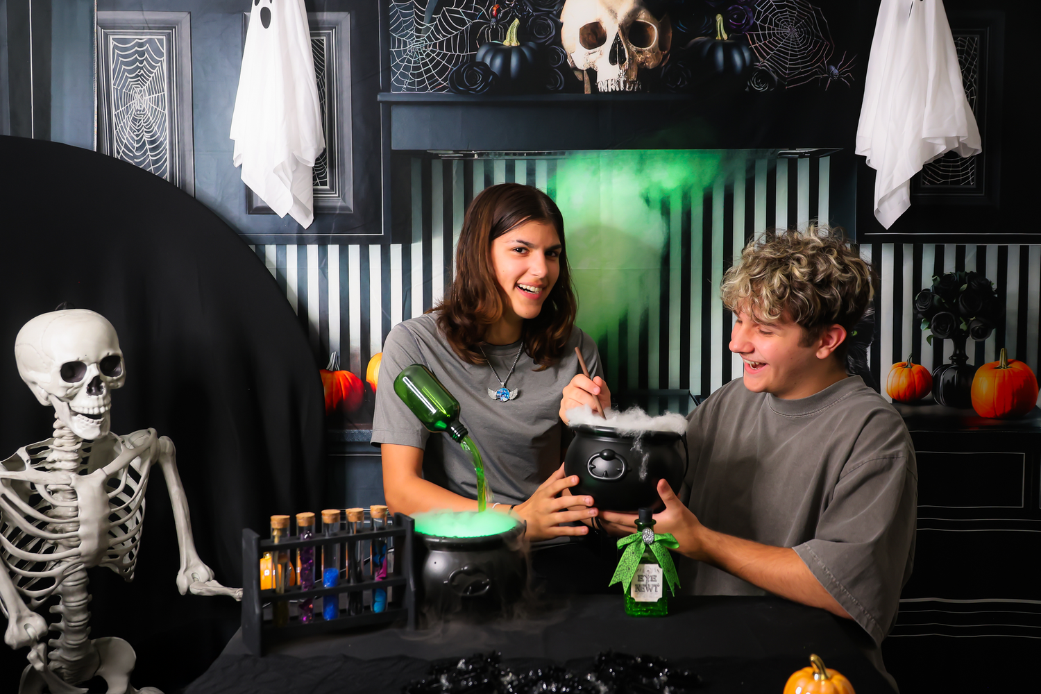 Two teens create a spooky Halloween potion with dry ice in a cauldron, surrounded by skeleton and ghost decorations.
