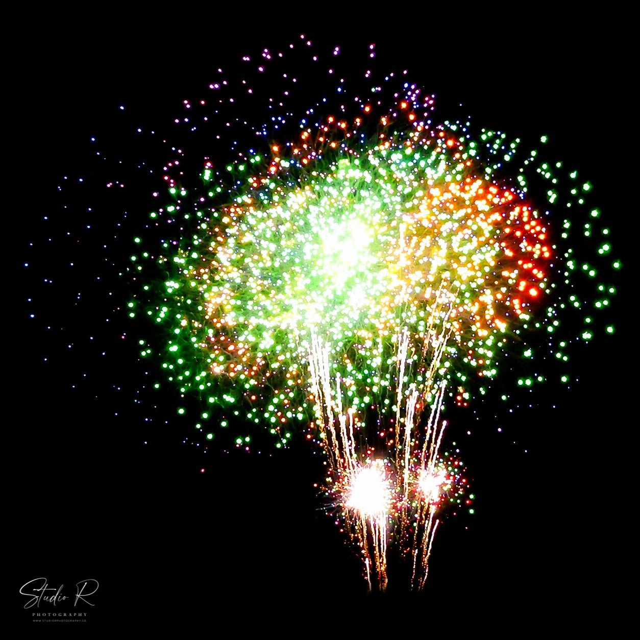 Colorful fireworks explosion with green, red, and orange sparks against a dark night sky.