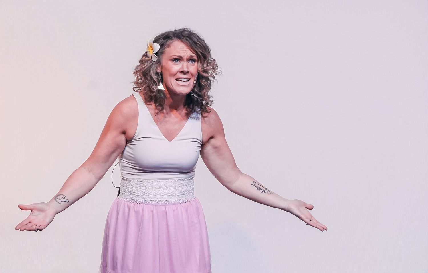 Woman in pink skirt and white top with tattoos, arms outstretched, flower in hair, performing on stage.