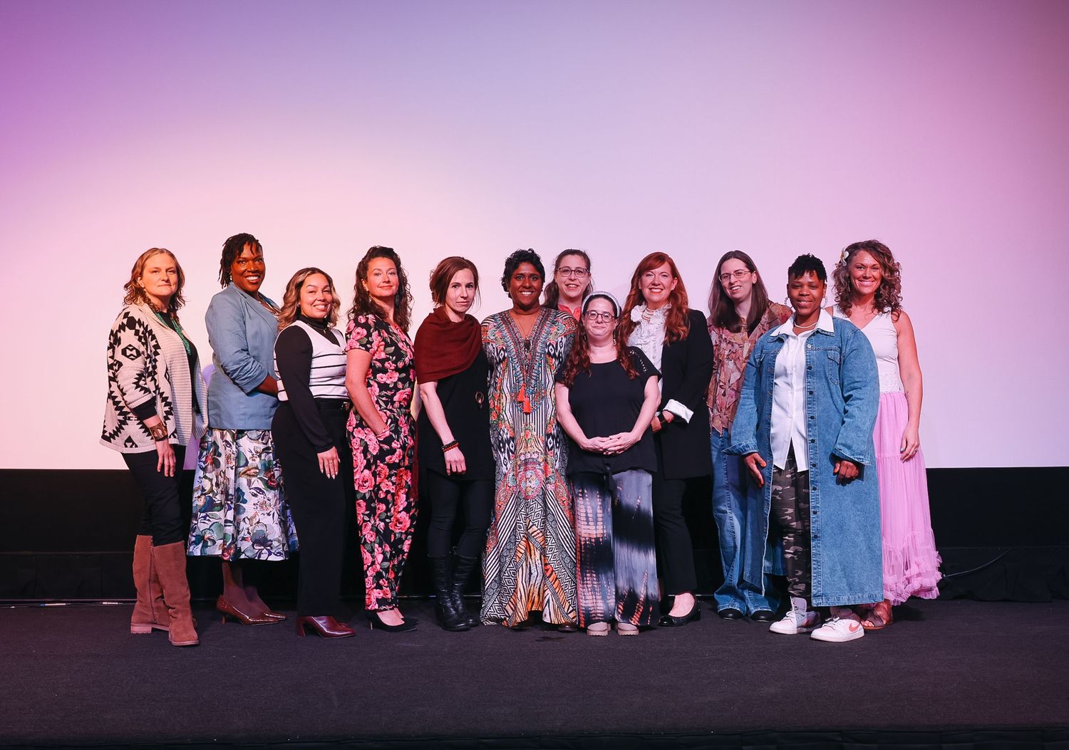 Group of diverse women posing together on stage at an event, smiling in front of a pink and purple backdrop.