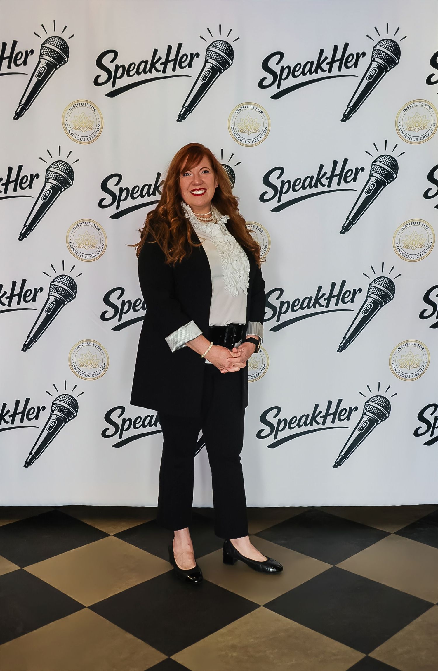 Woman in black blazer and pants poses at SpeakHer event backdrop with microphone logos.