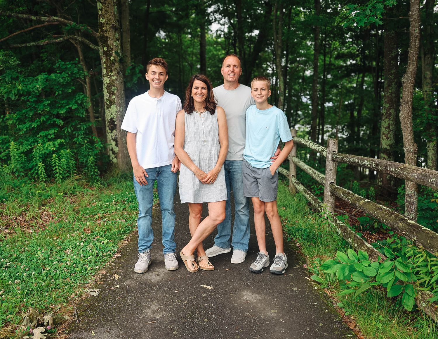 Family of four smiling on a scenic wooded path with a wooden fence in lush green surroundings.