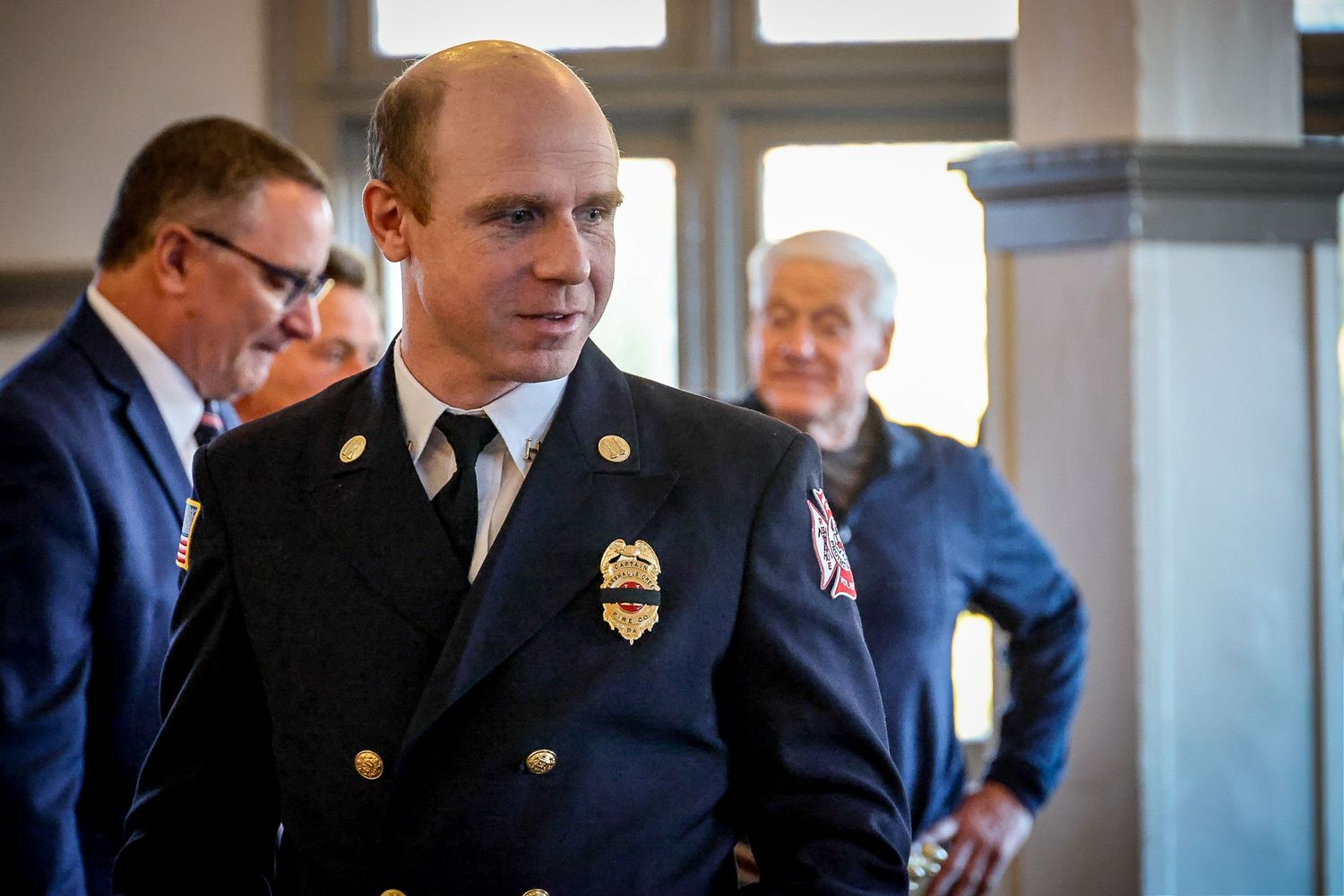 A firefighter in dress uniform with gold badge stands in a formal indoor setting with officials in background.