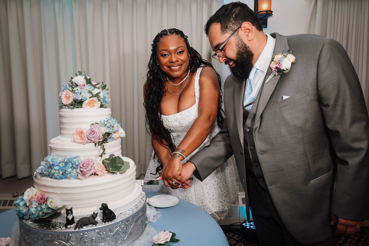 Bride and groom cutting their elegant four-tier white wedding cake decorated with colorful flowers at their reception.