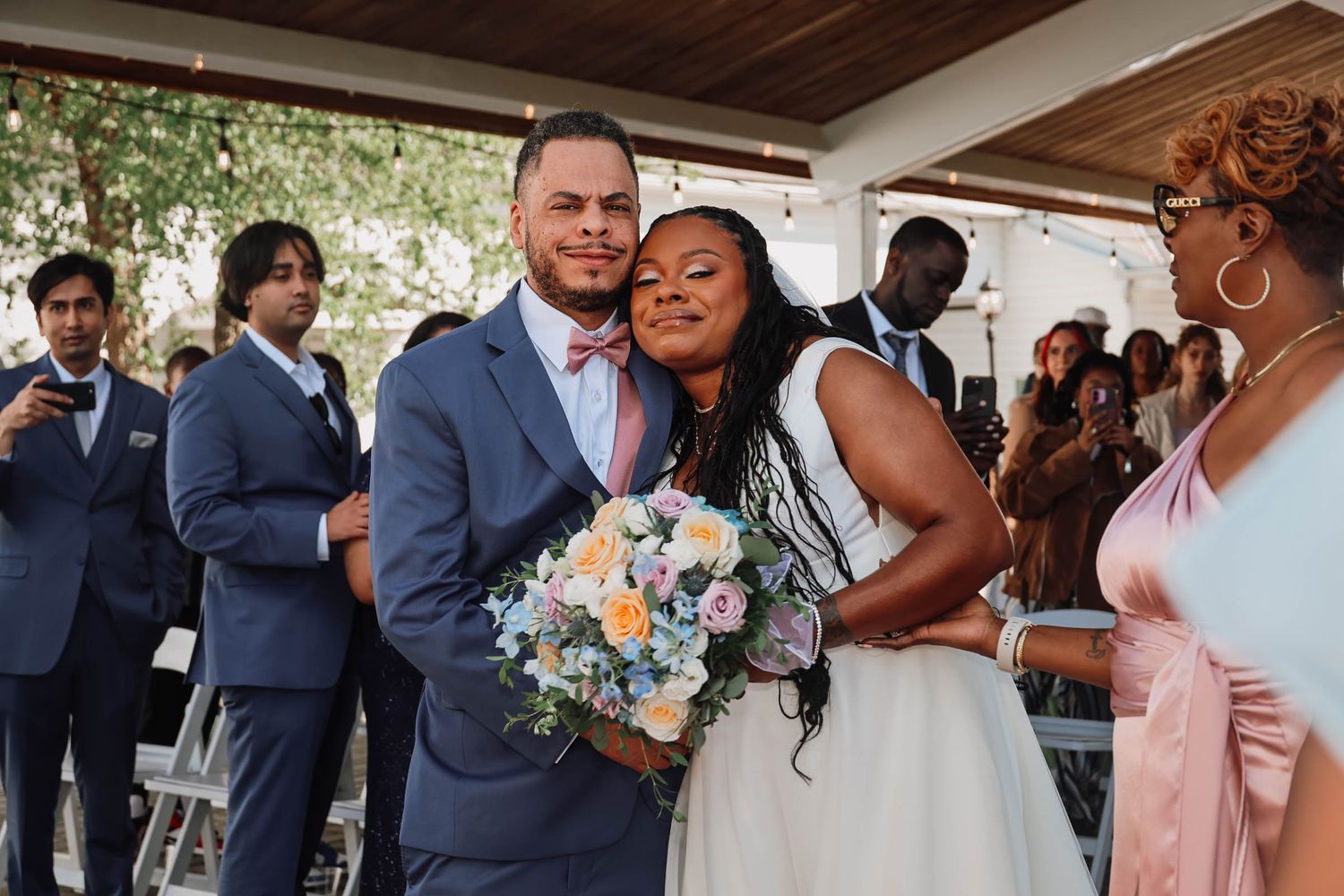 Smiling newlywed couple posing together after wedding ceremony, bride holding colorful bouquet, guests in background.
