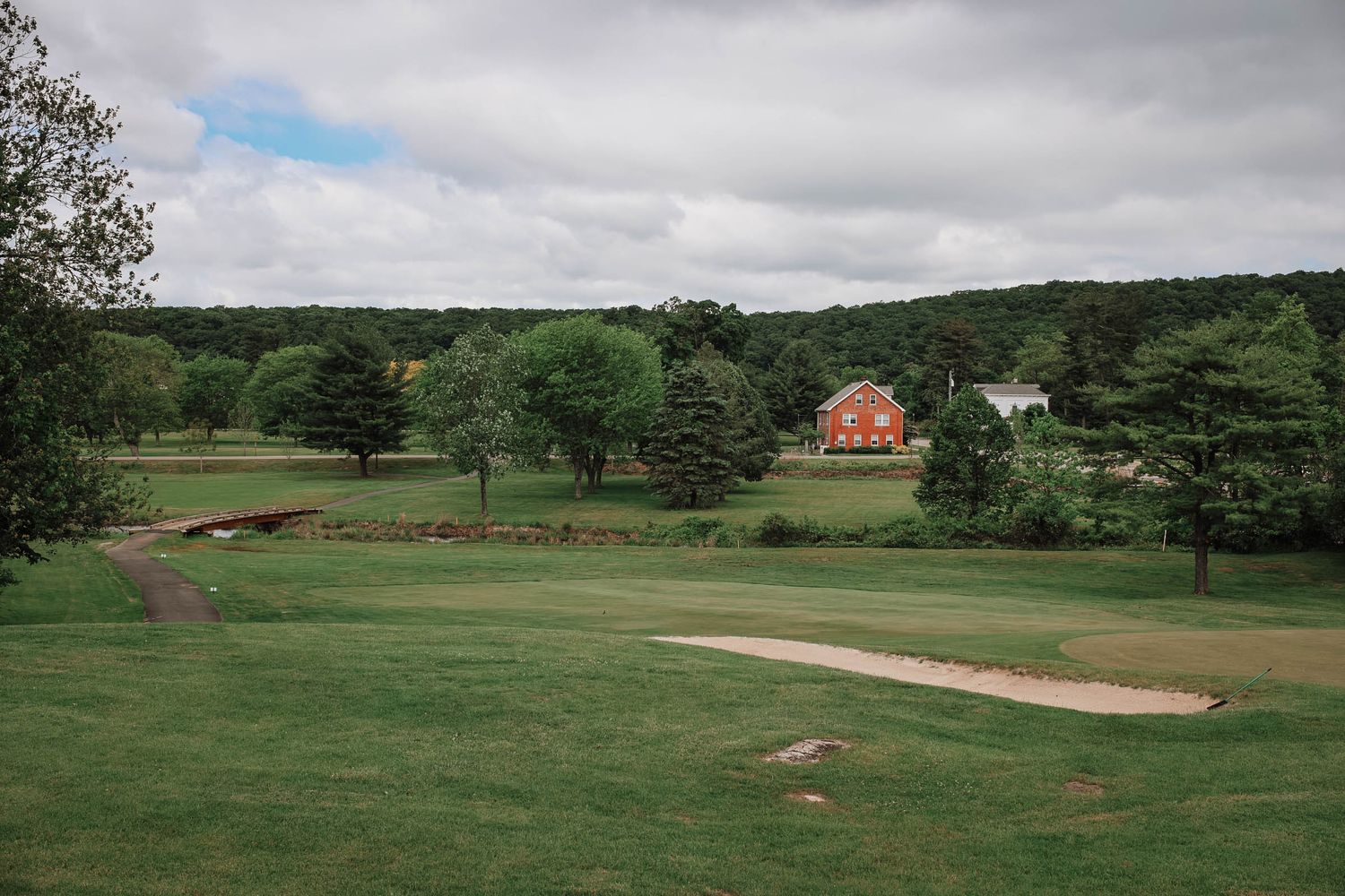 Lush green golf course with sand bunker, red barn, and tree-lined background under cloudy skies.