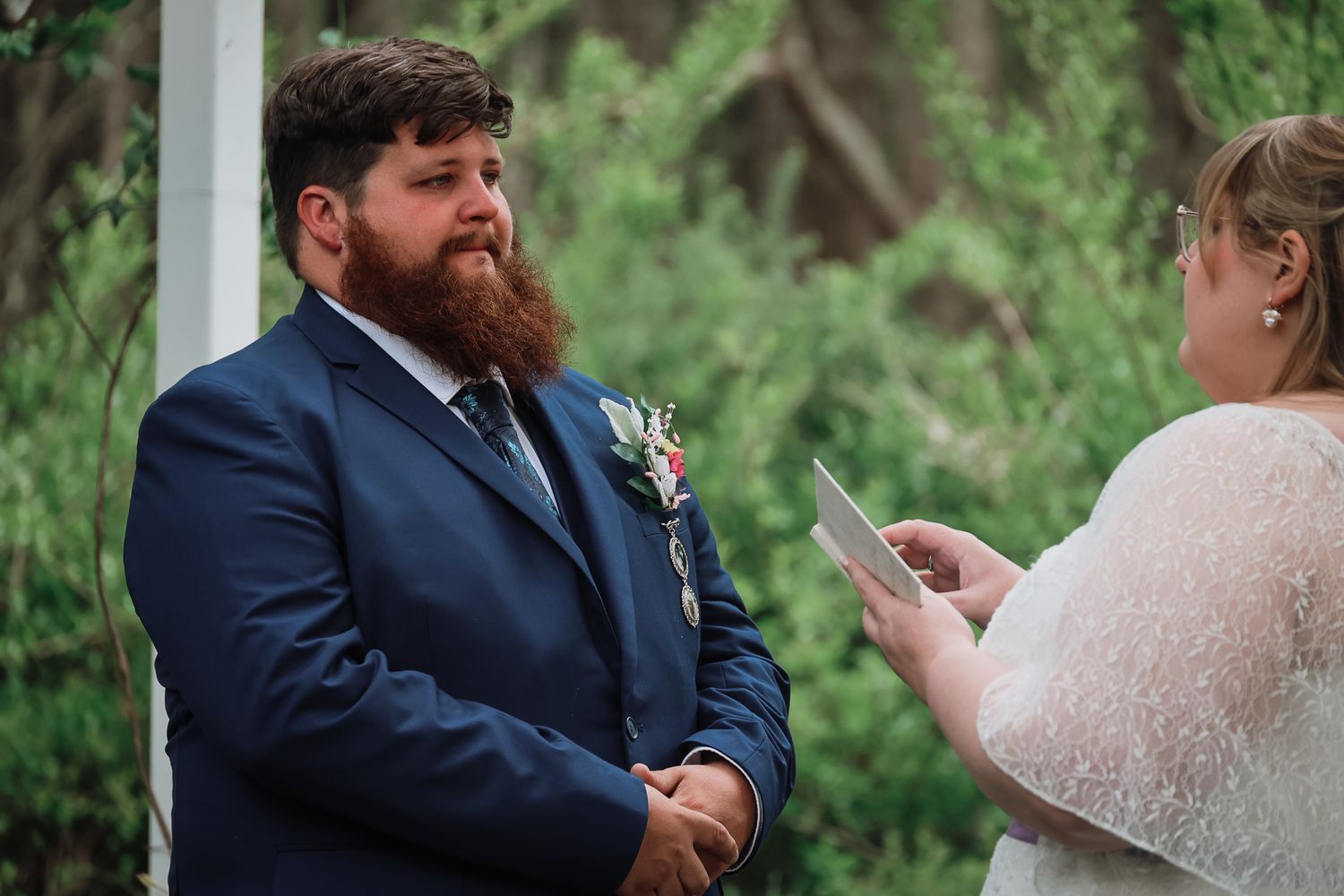 Groom in navy suit with boutonniere listens emotionally as bride reads wedding vows during outdoor ceremony.