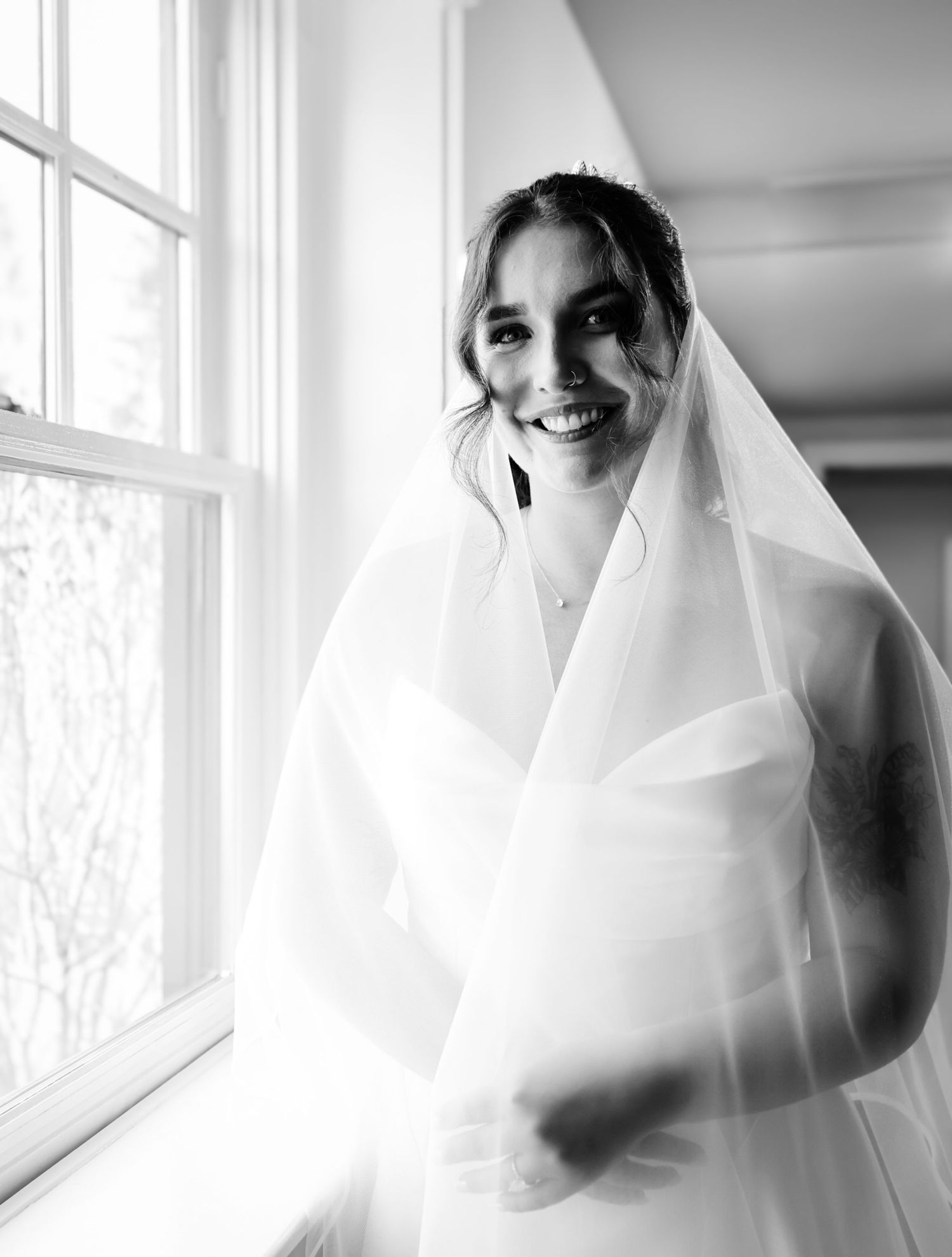 Black and white photo of a smiling bride in a wedding dress and veil standing by a window.