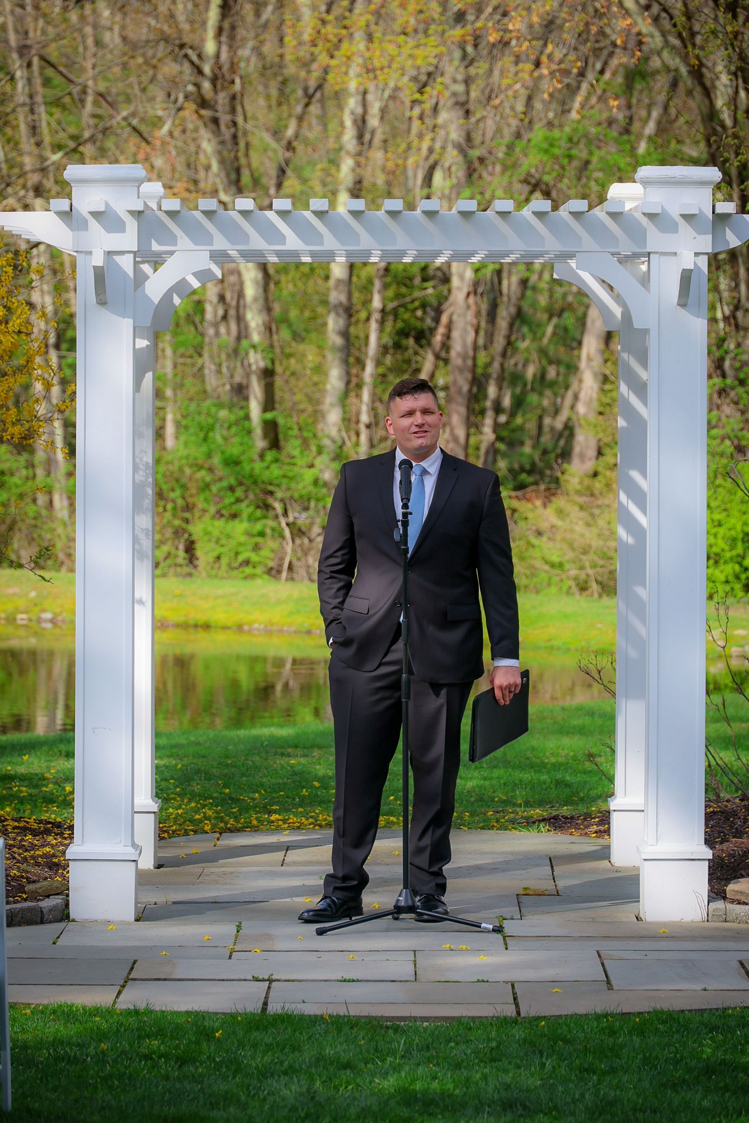 Man in dark suit stands under white pergola arch outdoors near pond and green trees.