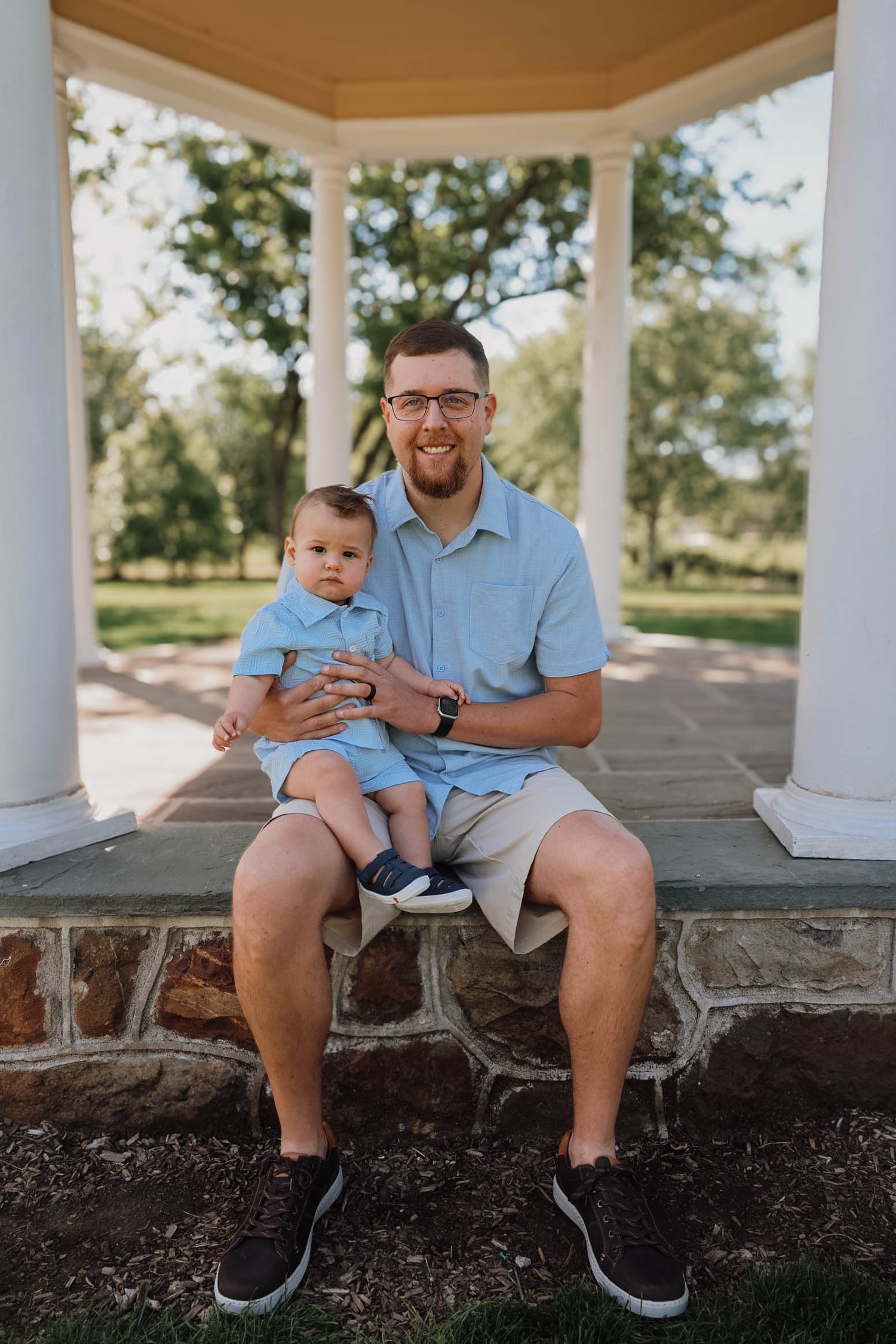 Man in glasses sits with baby under a white gazebo in a park, both wearing matching light blue outfits.