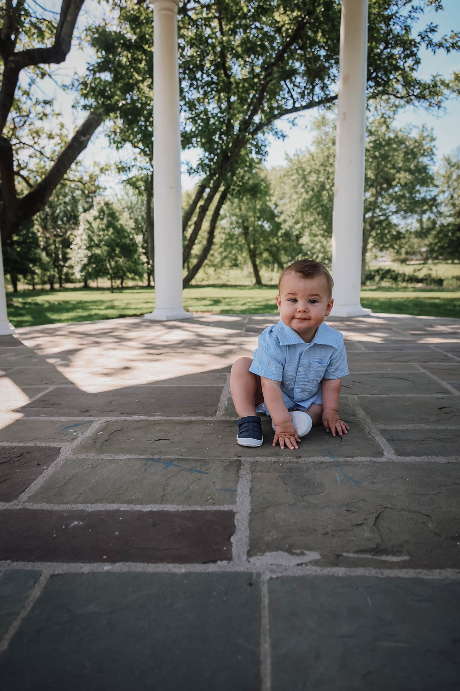 A baby boy in a blue romper sits on stone pavers near white columns in a lush green park.