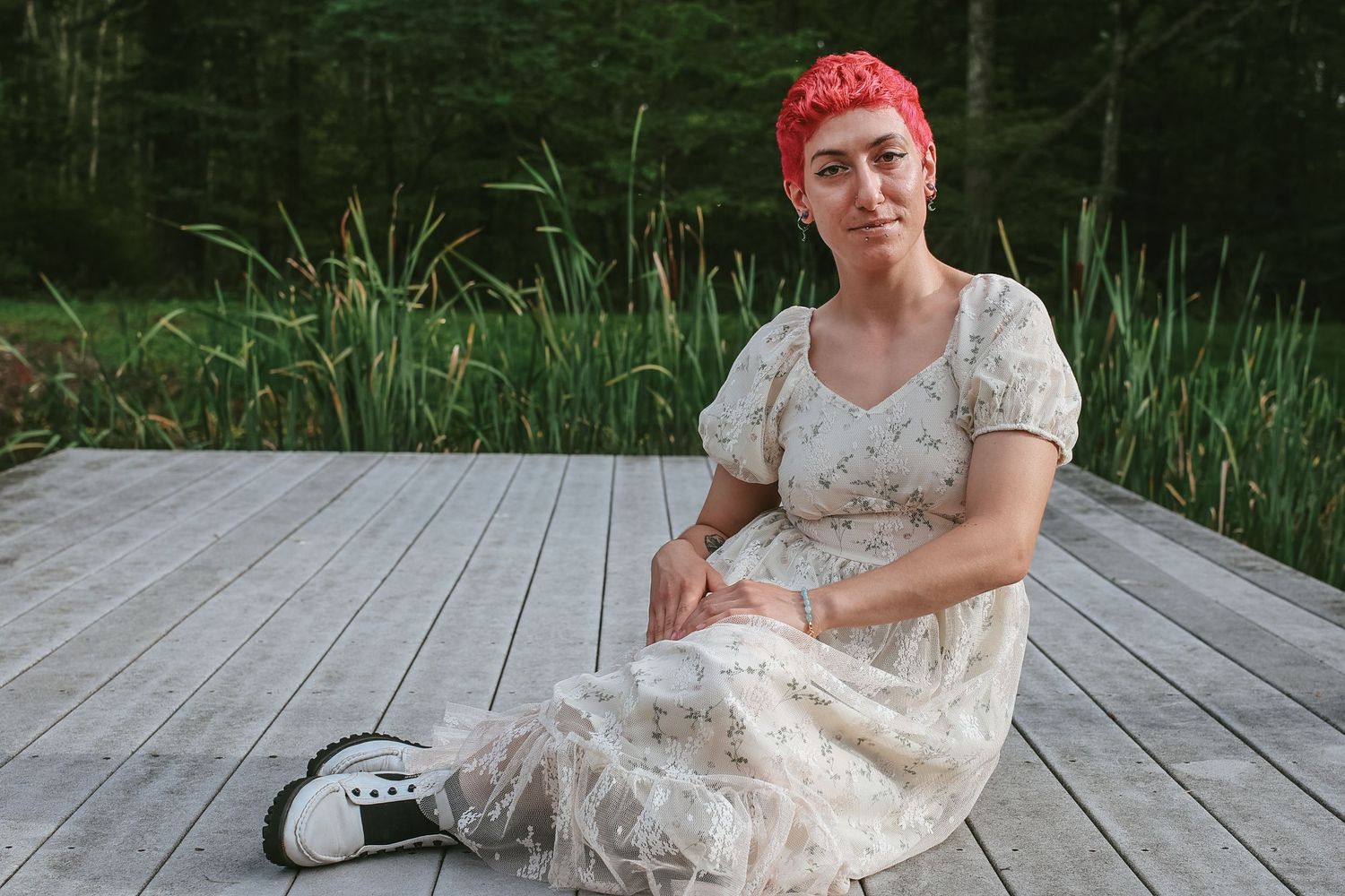Person with bright pink hair wearing a white lace dress sits on a wooden deck near tall grass and reeds.