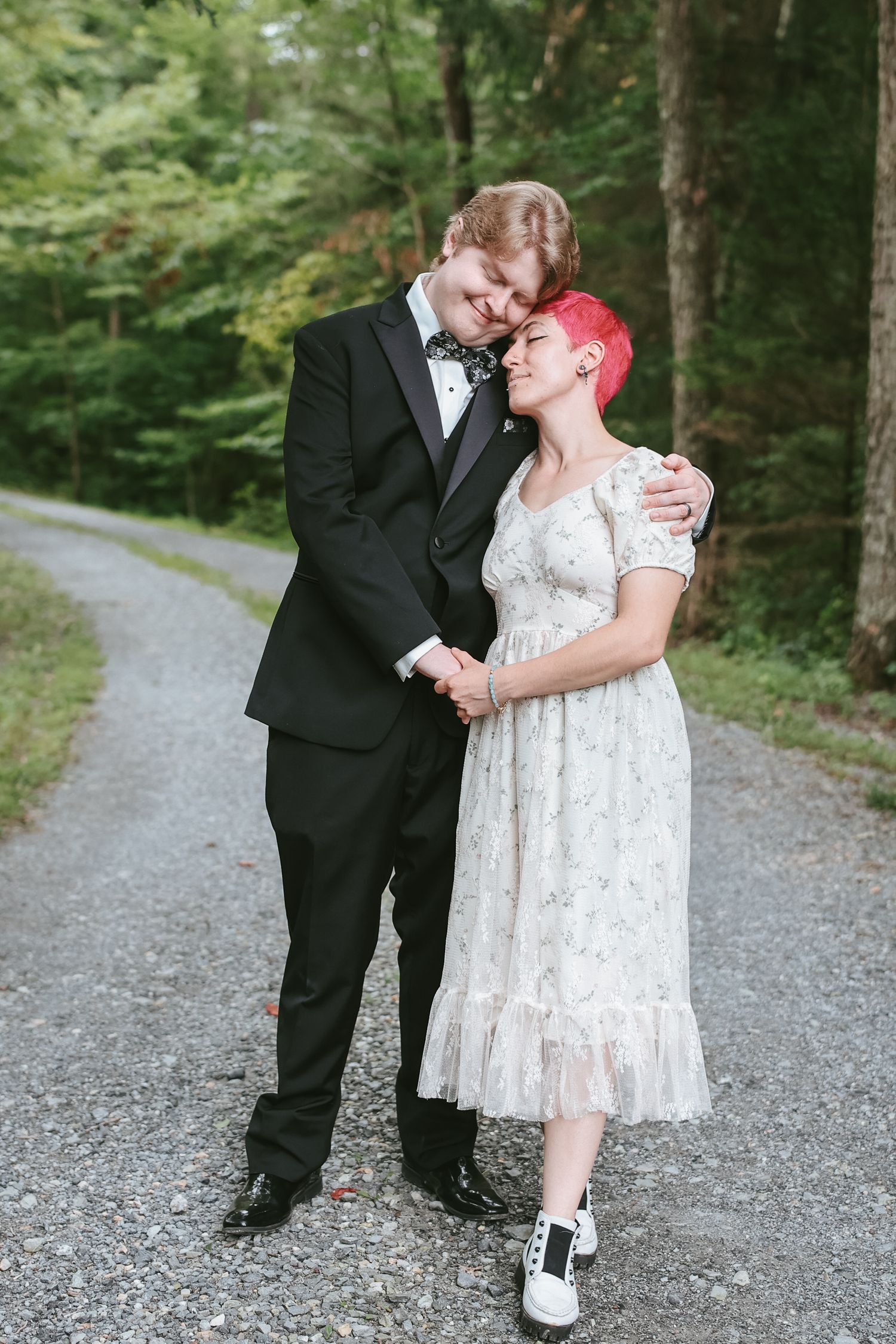 A couple shares a tender moment on a wooded gravel path while dressed in formal attire.