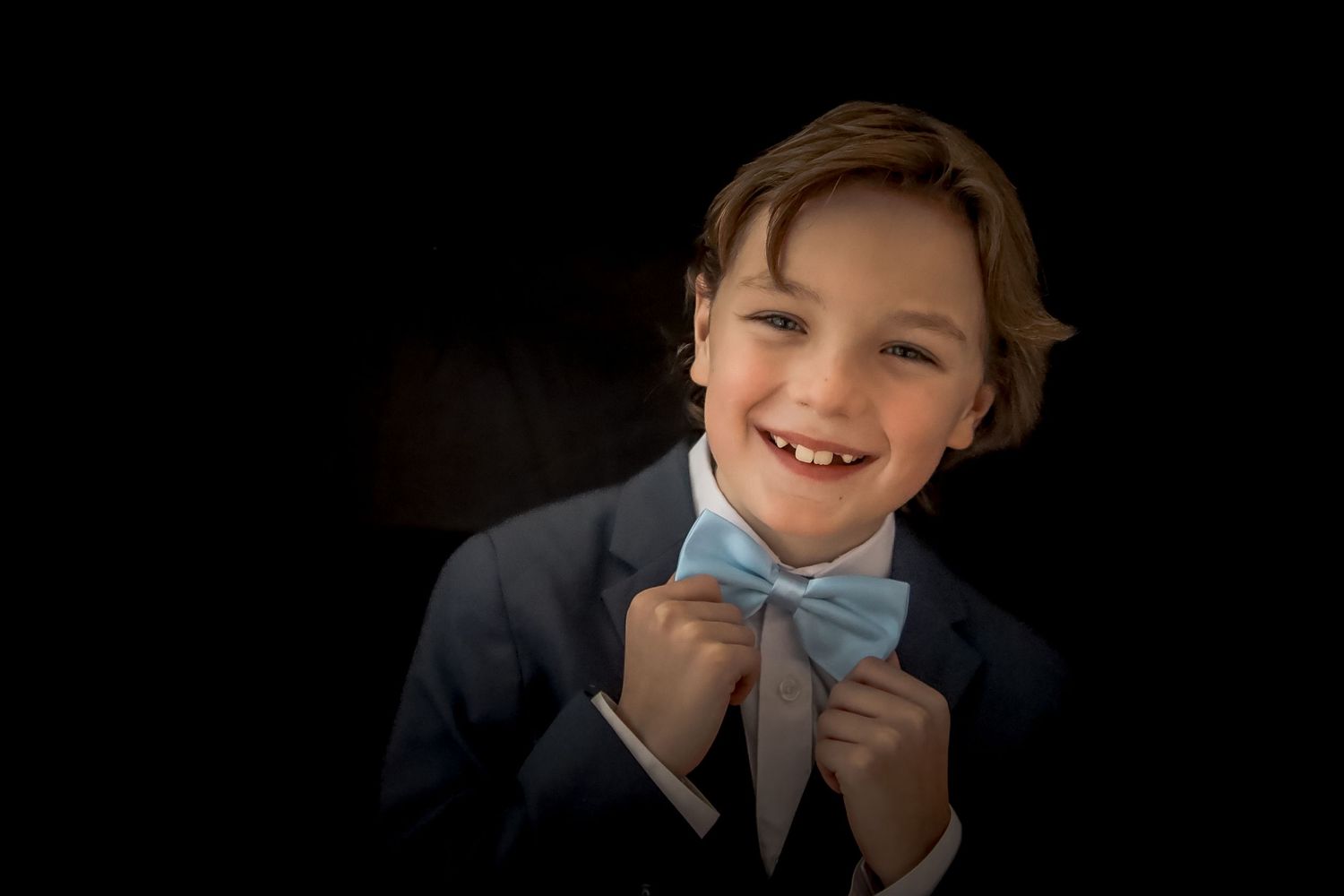 A child in a navy suit and light blue bowtie smiles warmly against a dark background.